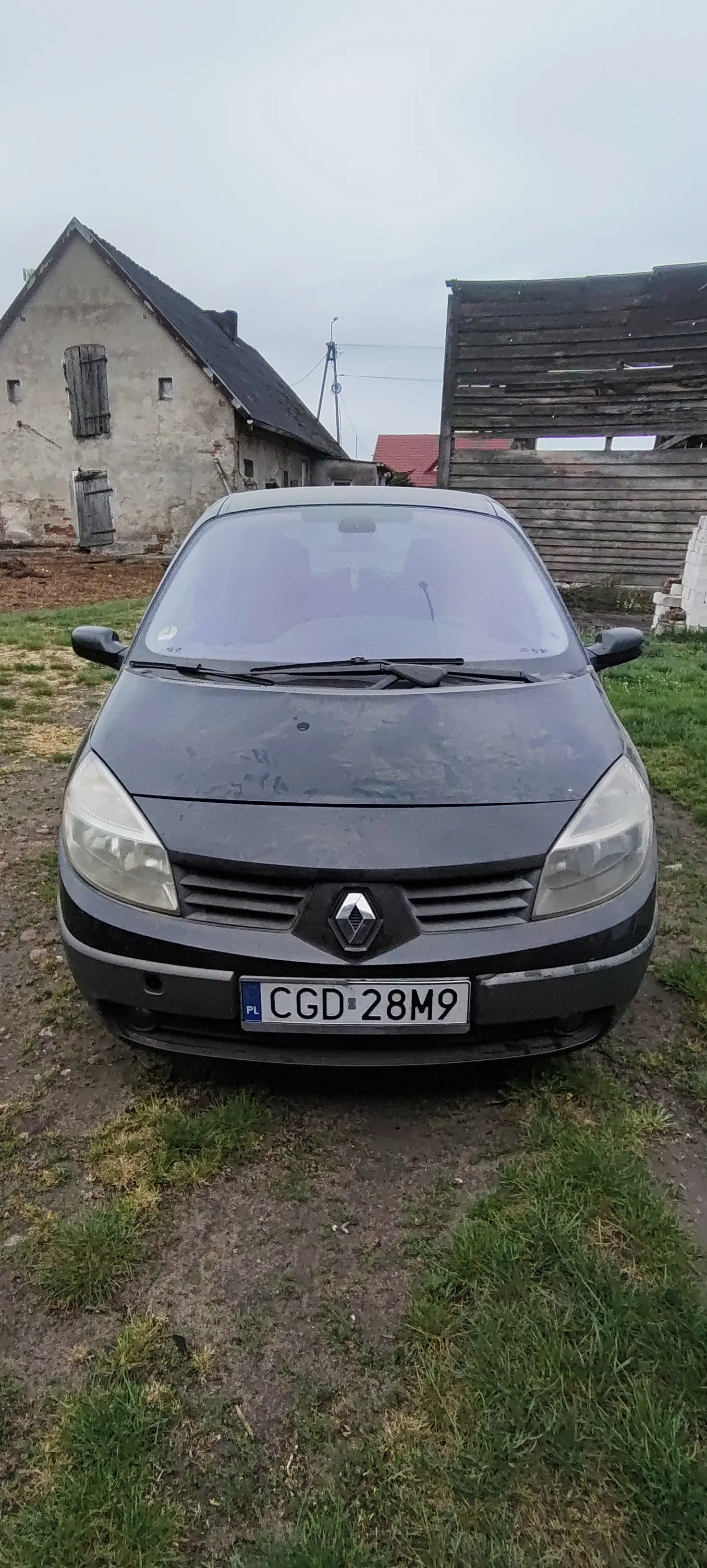 Szary samochód marki Renault z tablicą rejestracyjną CGD 28M9 (PL) zaparkowany na drodze ziemnej obok trawy. W tle stary budynek z ciemnym dachem, zamurowanymi oknami oraz drewnianą konstrukcją. Niebo jest pochmurne.