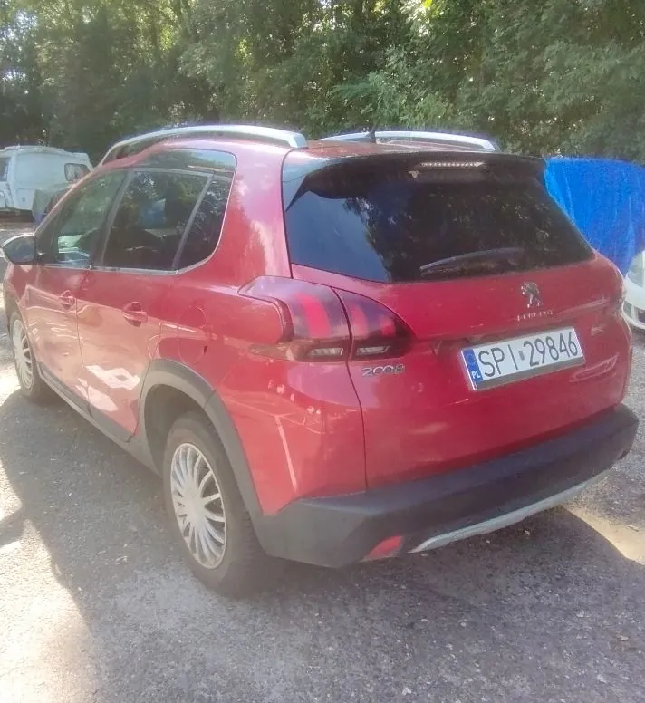 Czerwony samochód marki Peugeot 2008 zaparkowany na szutrowym podłożu. Widok z tyłu i z boku. Pojazd posiada polską tablicę rejestracyjną o numerze SPI 29846. W tle widoczne są zielone drzewa oraz fragment niebieskiego plandeki.