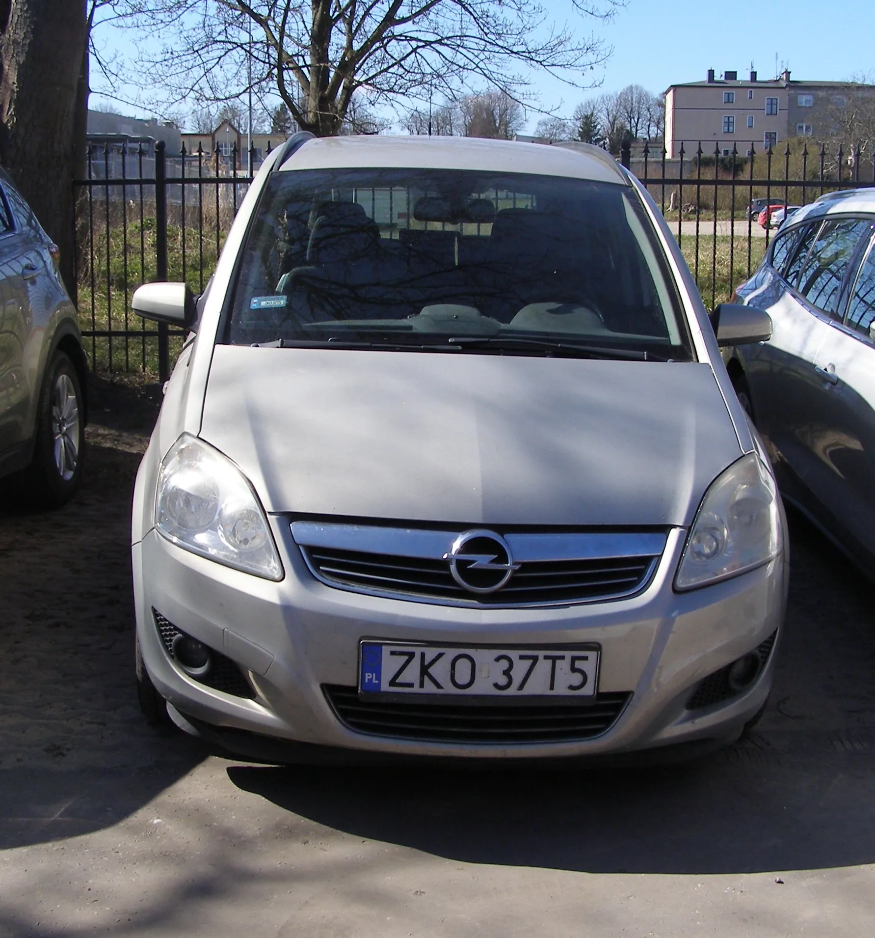 Srebrny samochód marki Opel z tablicą rejestracyjną ZKO 37T5 (PL) zaparkowany na otwartym placu. Po obu stronach widoczne są części innych pojazdów. W tle czarny metalowy płot, drzewa bez liści, budynki mieszkalne oraz jasne niebo. Na przedniej szybie samochodu znajduje się niebieski znaczek.