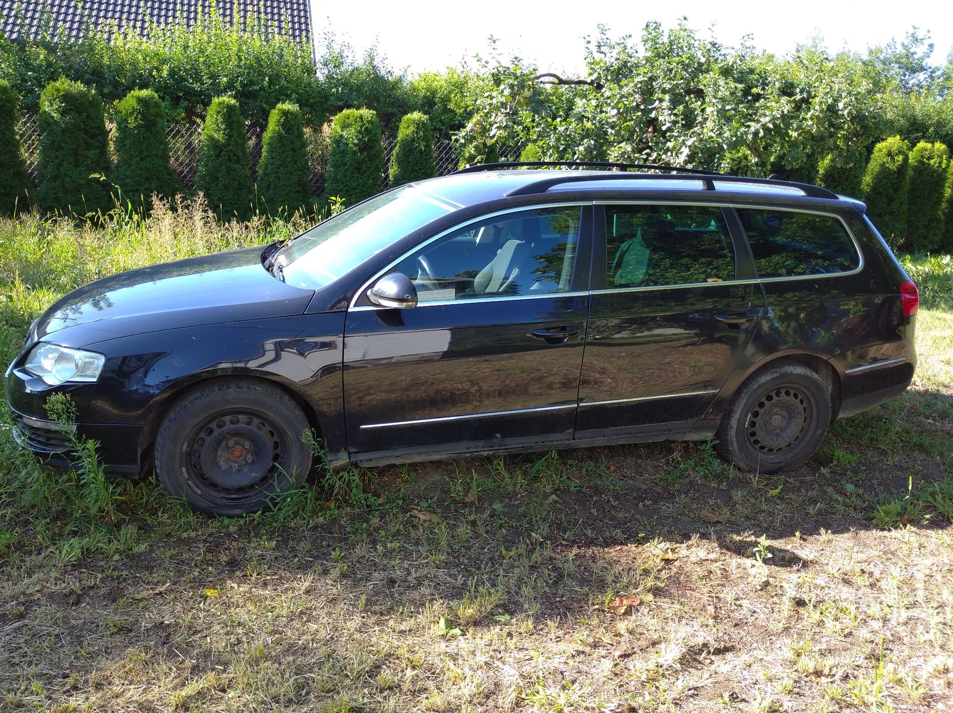 Czarny samochód typu kombi zaparkowany na trawiastym terenie. Pojazd jest widoczny z profilu, z lewej strony. W tle znajduje się gęsty żywopłot z zielonych krzewów oraz fragment dachu budynku z ciemną dachówką.