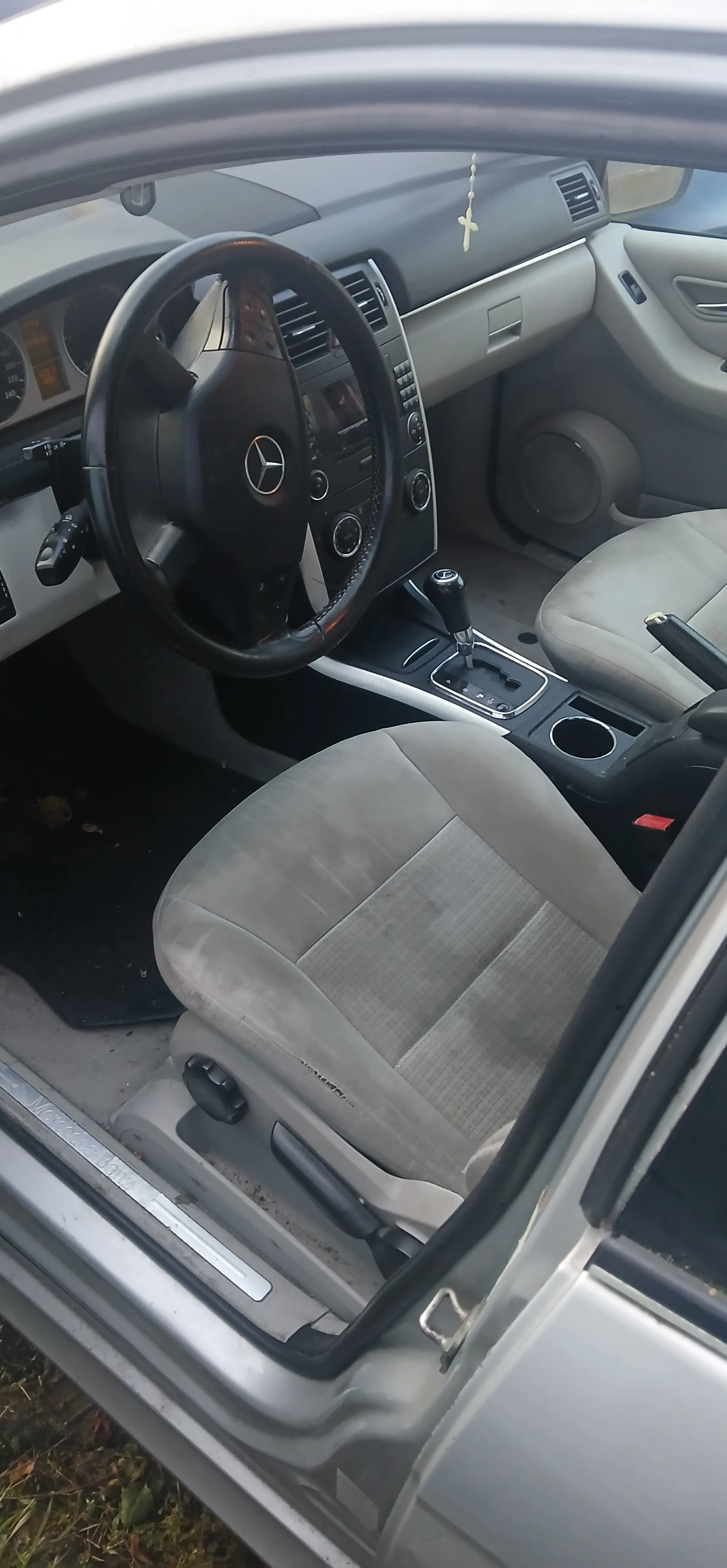 Wnętrze samochodu marki Mercedes-Benz z widocznym kierownicą z logo, szarymi siedzeniami, dźwignią zmiany biegów, deska rozdzielcza z przyciskami i wentylacją, wiszącym krzyżykiem na lusterku, otwartą drzwiami i fragmentem zewnętrznej obudowy pojazdu. Na podłodze widoczny czarny dywanik, a na zewnątrz fragment trawy.