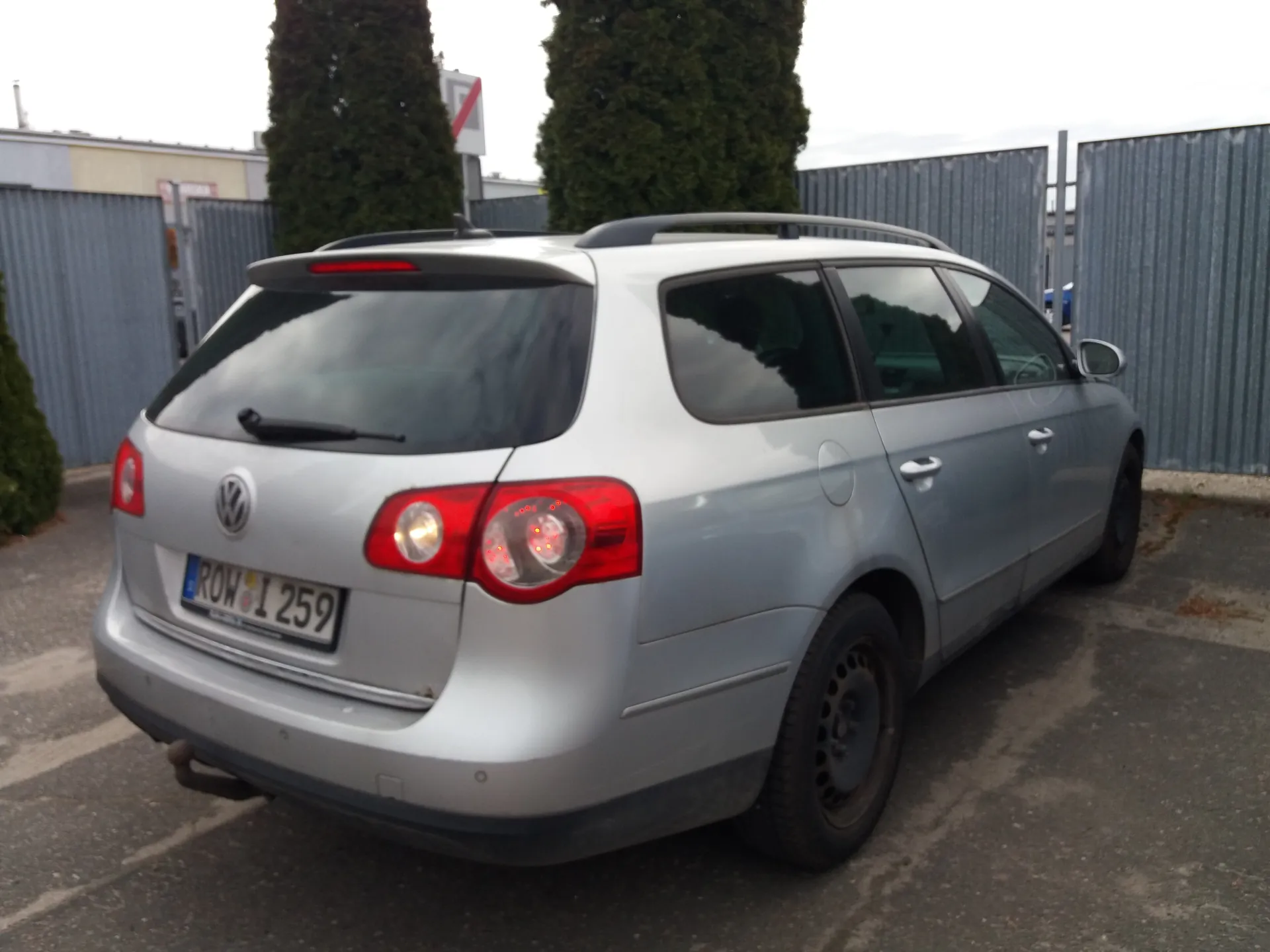 Srebrny samochód osobowy typu kombi marki Volkswagen zaparkowany na placu parkingowym. Widok od tyłu z widoczną tablicą rejestracyjną ROW 1259. W tle metalowe ogrodzenie, wysokie zielone drzewa i budynek z czerwonym znakiem. Niebo jest pochmurne.