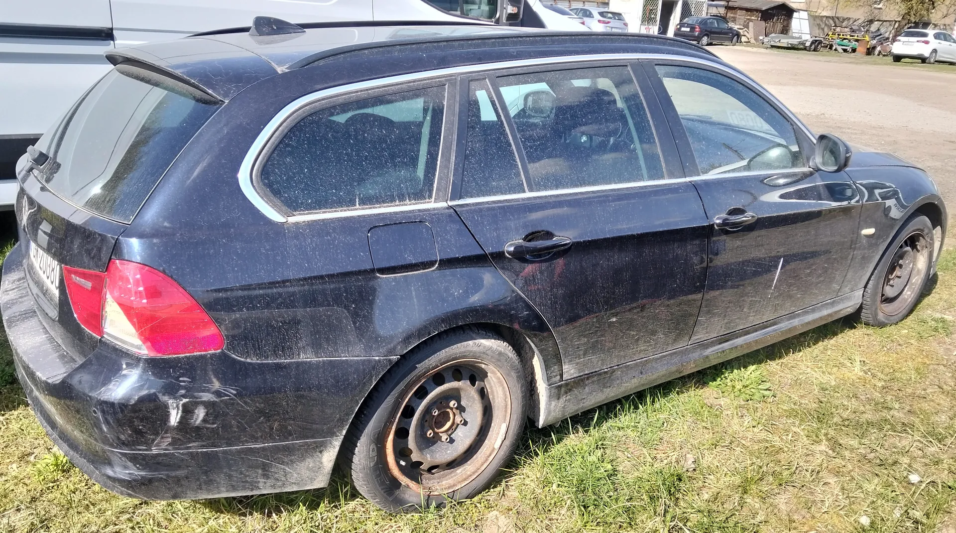 Czarny samochód typu kombi zaparkowany na trawie. Pojazd jest zabrudzony, z widocznymi śladami kurzu i zadrapaniami. Widoczne są czerwone światła tylne, tablica rejestracyjna z numerem WU2088L oraz felgi bez opon. W tle znajdują się inne pojazdy, budynek i narzędzia.
