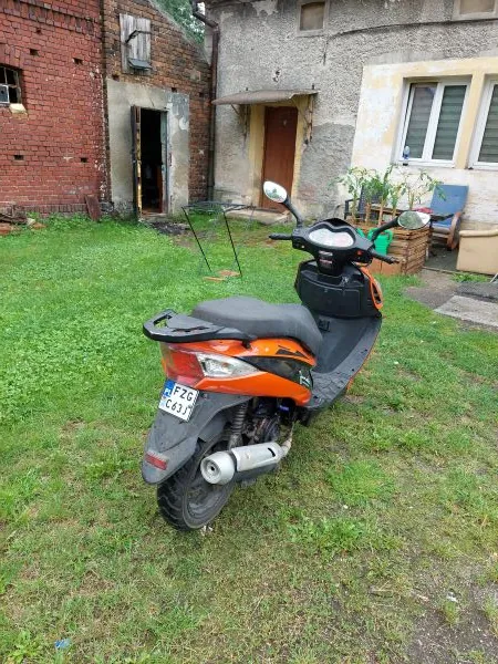 Pomarańczowo-czarny skuter zaparkowany na trawniku przed budynkiem. Skuter ma tablicę rejestracyjną o numerze PZG C63J. W tle widoczny jest fragment starego budynku z cegły oraz jasna ściana z oknem.