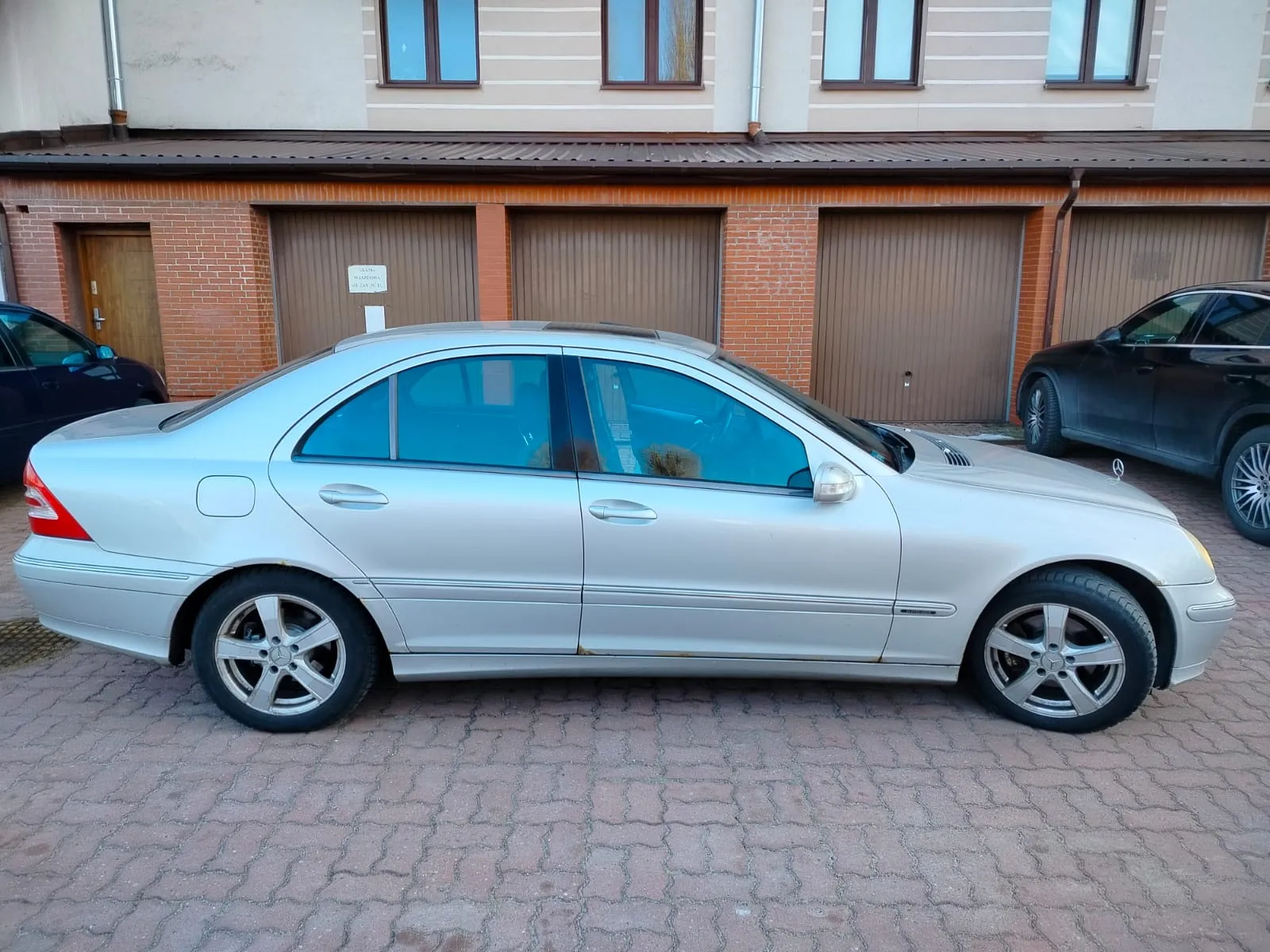 Srebrny sedan Mercedes-Benz z panoramicznym dachem, przyciemnianymi szybami i aluminiowymi felgami zaparkowany na brukowanej powierzchni. W tle ceglany budynek z brązowymi garażami i oknami. Po bokach czarne samochody. Na progu drzwi widoczne ślady rdzy.