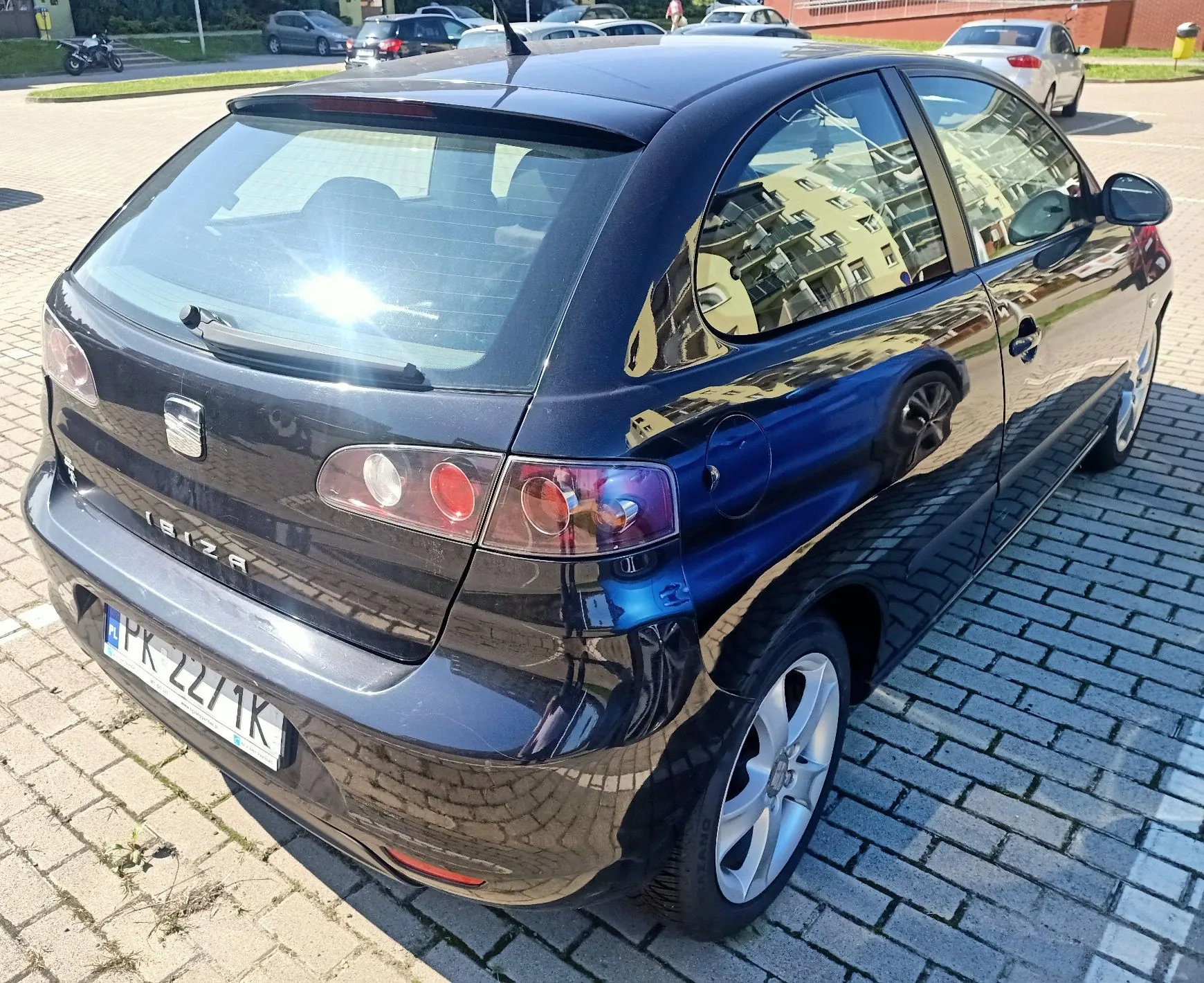 Czarny samochód marki Seat Ibiza zaparkowany na kostce brukowej. Widok z tyłu i z boku pojazdu. Samochód ma tablicę rejestracyjną o numerze PK 2271K. W tle widoczne są inne zaparkowane samochody oraz budynki mieszkalne odbijające się w karoserii i szybach auta.