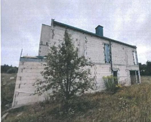 Budynek z białych bloków betonowych o dwóch kondygnacjach, z niebieskim kominem na dachu. Przed budynkiem rośnie drzewo, a wokół trawa i krzewy. Budynek ma kilka okien z niebieskimi ramami oraz drzwi. Tło stanowi pochmurne niebo i otaczający teren z roślinnością. Budynek wydaje się być nieco pochylony w lewo.