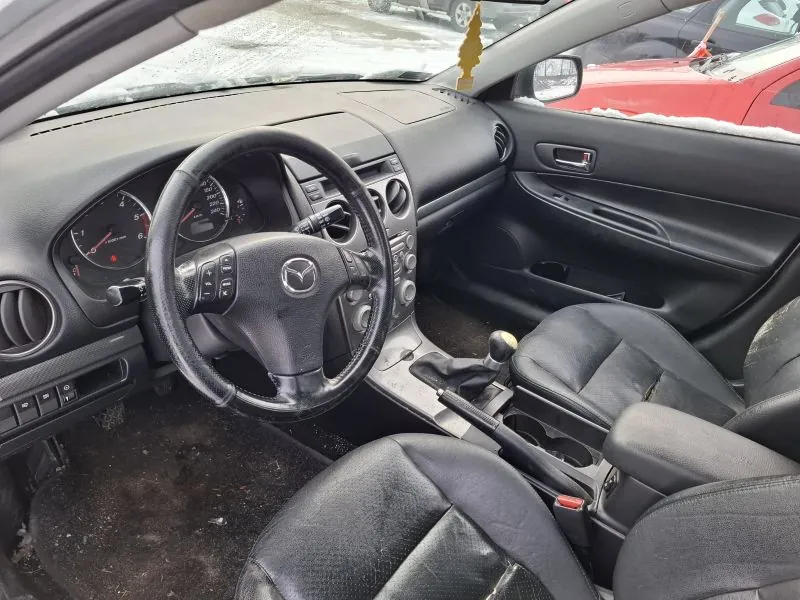 Wnętrze samochodu marki Mazda z widocznym kierownicą z logo, czarną tapicerką, deską rozdzielczą z wskaźnikami prędkości i obrotomierzem, dźwignią zmiany biegów oraz czarnymi skórzonymi fotelami. Podłoga jest zabrudzona, a przez przednią szybę widać śnieg na ziemi i czerwony samochód na zewnątrz. Drzwi pasażerskie z uchwytem i sterowaniem szybą są widoczne po prawej stronie.