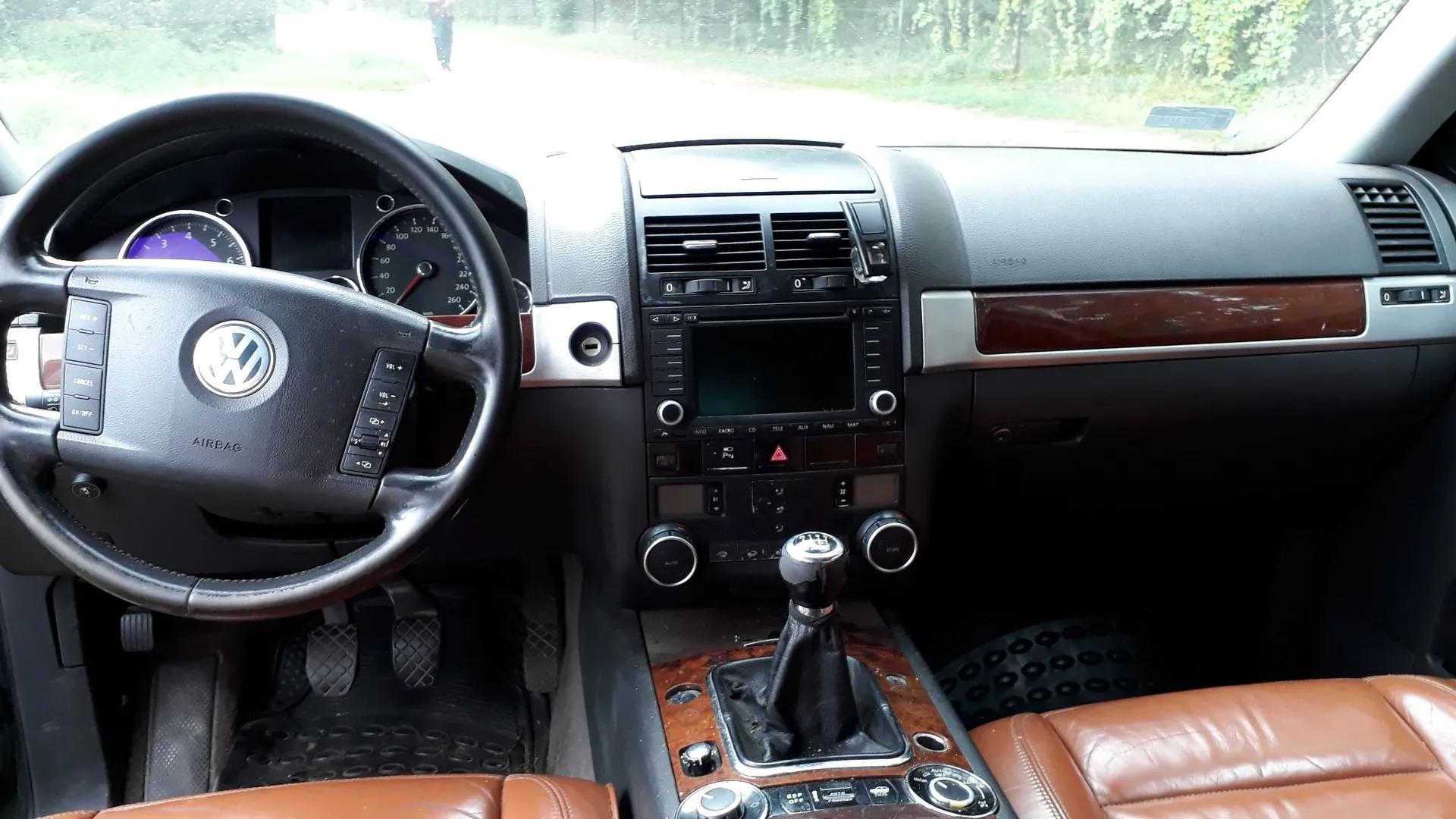 Wnętrze samochodu marki Volkswagen, widok z perspektywy kierowcy na deskę rozdzielczą. Po lewej stronie znajduje się czarna kierownica z logo VW i napisem AIRBAG. Za nią widoczne są zegary analogowe i cyfrowy wyświetlacz. W centrum znajduje się konsola środkowa z nawiewami, ekranem multimedialnym, przyciskami sterowania klimatyzacją oraz lewarkiem zmiany biegów. Po prawej stronie widać fragment deski rozdzielczej z drewnianą wstawką. Fotele są wykonane z brązowej skóry.
