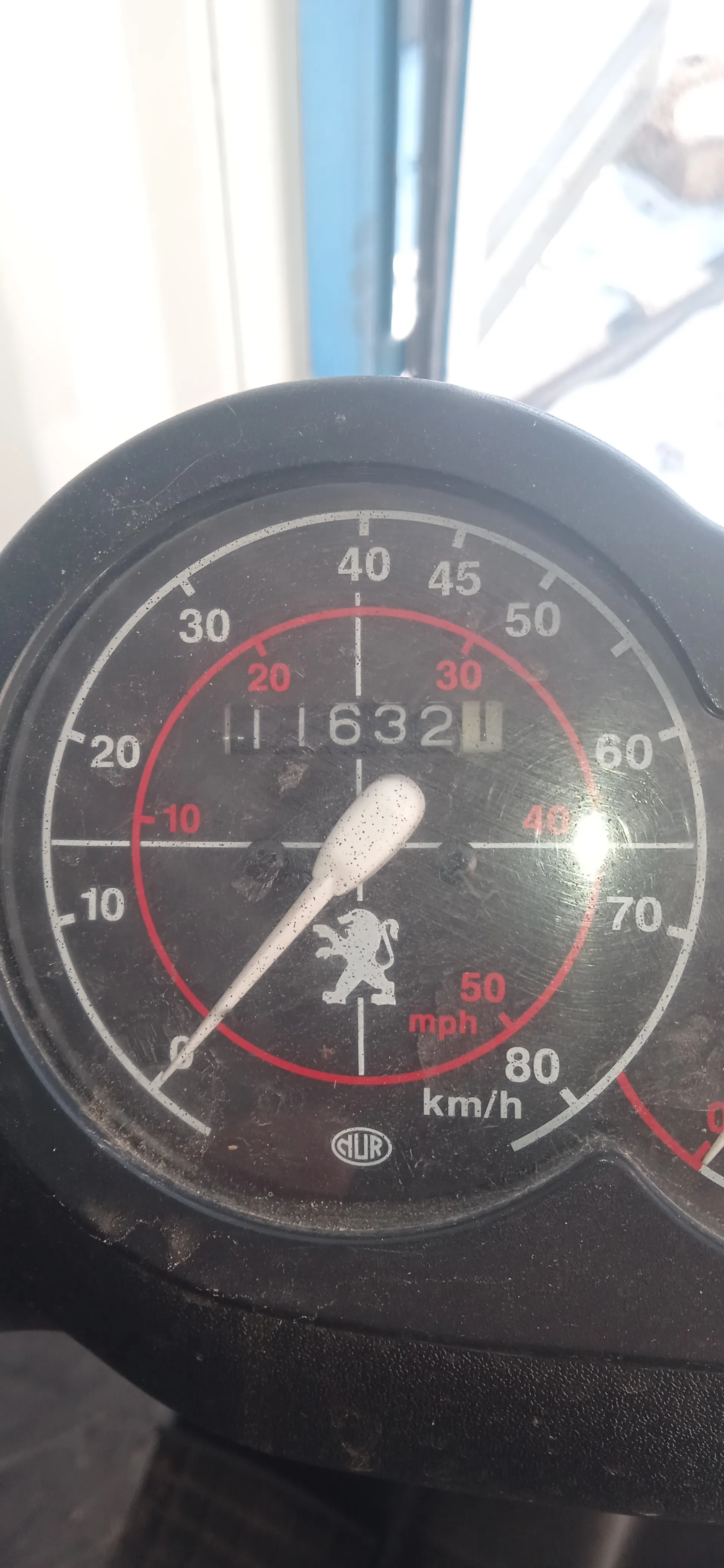 Zbliżenie na czarny licznik prędkości z białymi i czerwonymi oznaczeniami. Wskazówka wskazuje na wartość bliską 0. Na tarczy widoczne są skale: zewnętrzna w km/h (0-80) i wewnętrzna w mph (0-50). W centrum znajduje się biały symbol lwa oraz cyfry 11632 na wyświetlaczu przebiegu. W dolnej części tarczy widnieje napis 'CLUR' oraz oznaczenia 'km/h' i 'mph'. Tło jest rozmyte, z widocznymi elementami okna i niebieskim pionowym pasem.