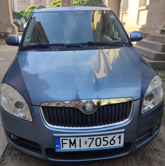 Widok z przodu na niebieski samochód marki Skoda zaparkowany na ulicy. Na masce i przedniej szybie widoczne są odbicia budynków. Samochód posiada polską tablicę rejestracyjną o numerze FMI 70561. Poniżej tablicy znajduje się niebieski pasek z napisem 'autokomismarek.pl Międzyrzecz ul. Kazimierza Wielkiego tel. 606 639 709'.