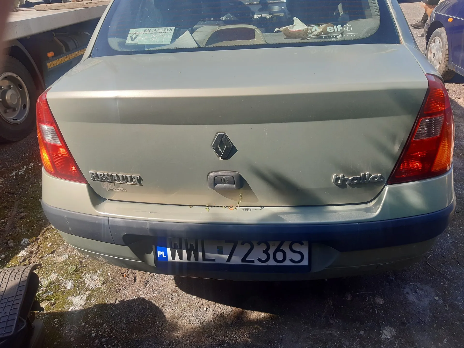Tylny widok srebrno-zielonego samochodu marki Renault Thalia. Na klapie bagażnika widoczne jest logo Renault oraz napisy 'RENAULT' po lewej i 'thalia' po prawej stronie. Samochód posiada polską tablicę rejestracyjną o numerze WWL 7236S. W tylnej szybie widoczne są naklejki, w tym jedna z napisem 'KULANCZYK' oraz 'elf'.