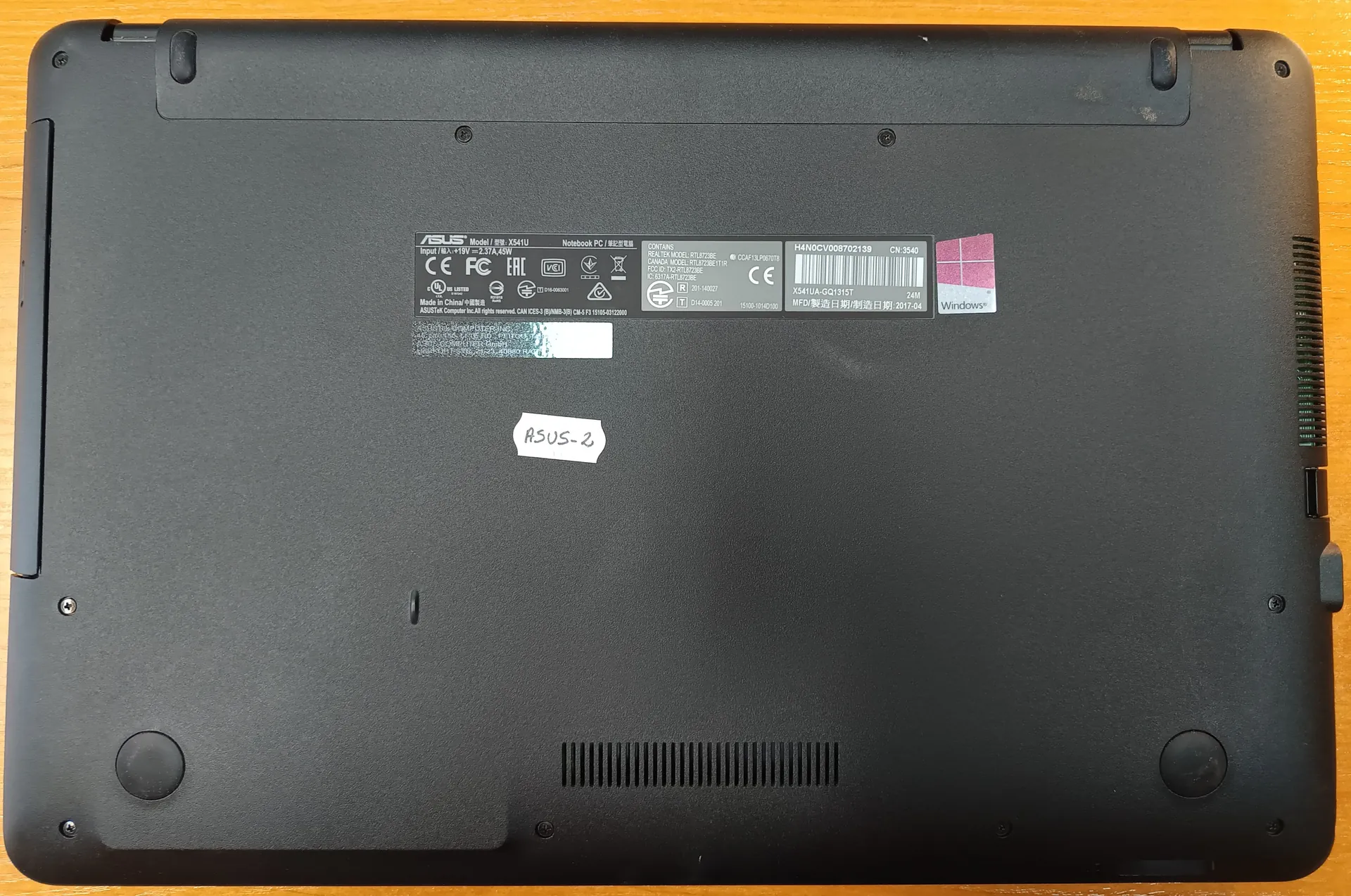 Czarna obudowa spodu laptopa marki ASUS model X541U na drewnianym tle. Widoczne etykiety z oznaczeniami: ASUS, CE, FC, EAC, Windows, kod kreskowy, numer seryjny H4NOCV008702139, data produkcji 2017-04 oraz ręcznie napisany naklejony napis ASUS-2. Na obudowie znajdują się również otwory wentylacyjne, śruby i okrągłe podstawki.