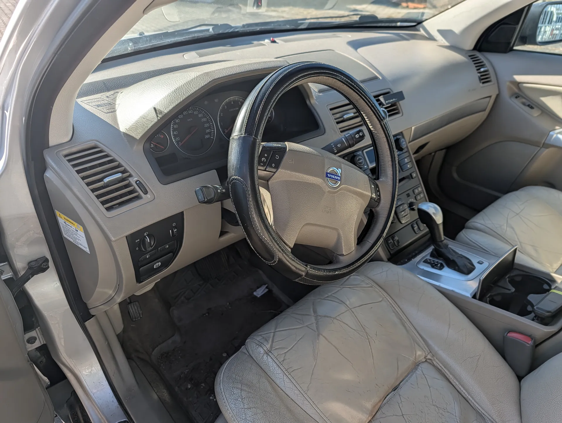 Wnętrze samochodu Volvo z kierownicą z logo VOLVO, deską rozdzielczą z licznikami prędkości i obrotomierzem, jasnobrązowymi usłojonymi siedzeniami, dźwignią zmiany biegów, otwartymi drzwiami kierowcy oraz brudnymi dywanami podłogowymi. Wnętrze w kolorze beżowym, widoczne również wentylacyjne otwory i elementy sterujące na desce rozdzielczej.