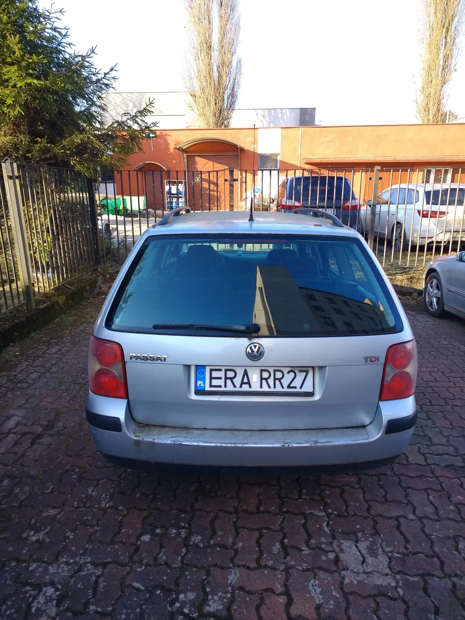 Srebrny samochód Volkswagen Passat TDI z tablicą rejestracyjną ERA RR27 (PL) zaparkowany na brukowanej powierzchni. W tle pomarańczowy budynek z bramą, metalowe ogrodzenie, drzewa oraz inne zaparkowane pojazdy. Widoczne są czerwone światła tylnego oraz napisy 'PASSAT' i 'TDI' na bagażniku.
