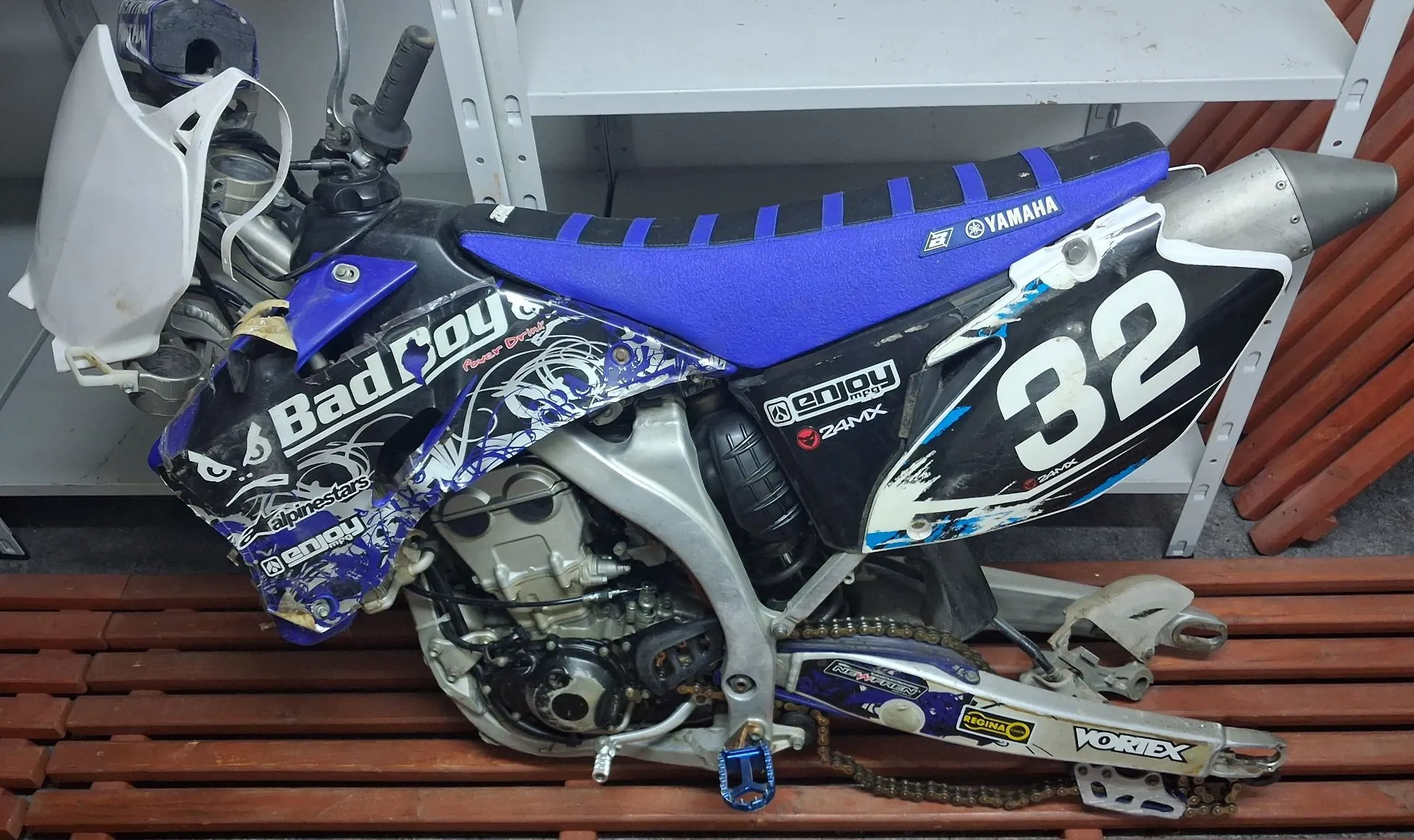 Motocykl crossowy w kolorach niebieskim i białym, ustawiony bokiem. Na boku motocykla widnieje duży biały numer '32' na czarnym tle oraz napisy 'Bad Boy', 'Yamaha', 'Alpinestars' i 'Enjoy'. Siedzisko jest w intensywnym niebieskim kolorze. Motocykl stoi na drewnianej kratownicy, a w tle widoczny jest biały regał.