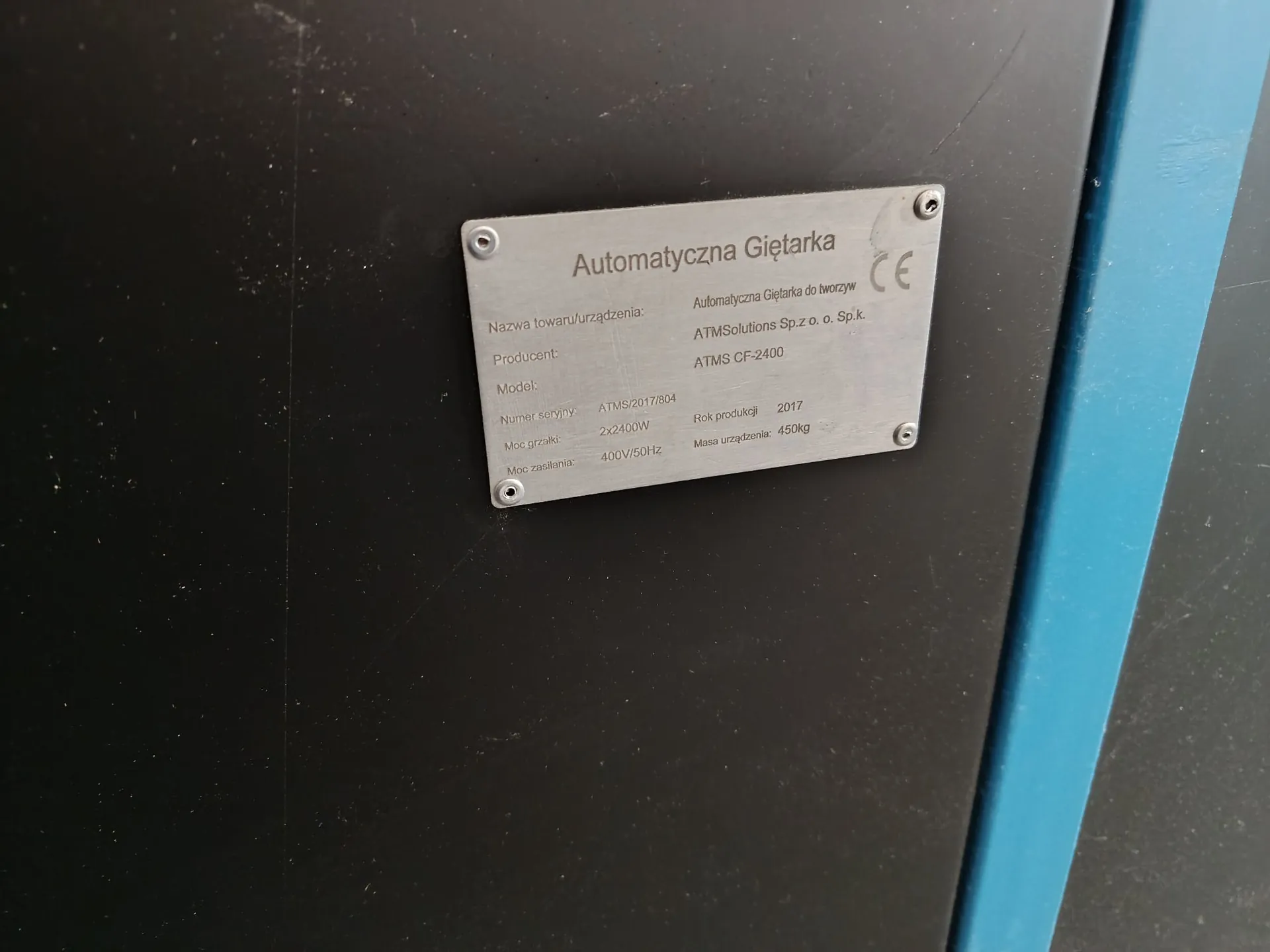Srebrna tabliczka znamionowa przymocowana do czarnej powierzchni za pomocą czterech śrub. Na tabliczce znajduje się napis: 'Automatyczna Giętarka'. Poniżej widnieją dane techniczne: Nazwa towaru/urządzenia: Automatyczna Giętarka do tworzyw; Producent: ATMSolutions Sp.z o. o. Sp.k.; Model: ATMS CF-2400; Numer seryjny: ATMS/2017/804; Rok produkcji: 2017; Moc grzałki: 2x2400W; Masa urządzenia: 450kg; Moc zasilania: 400V/50Hz. W prawym górnym rogu widnieje znak CE.