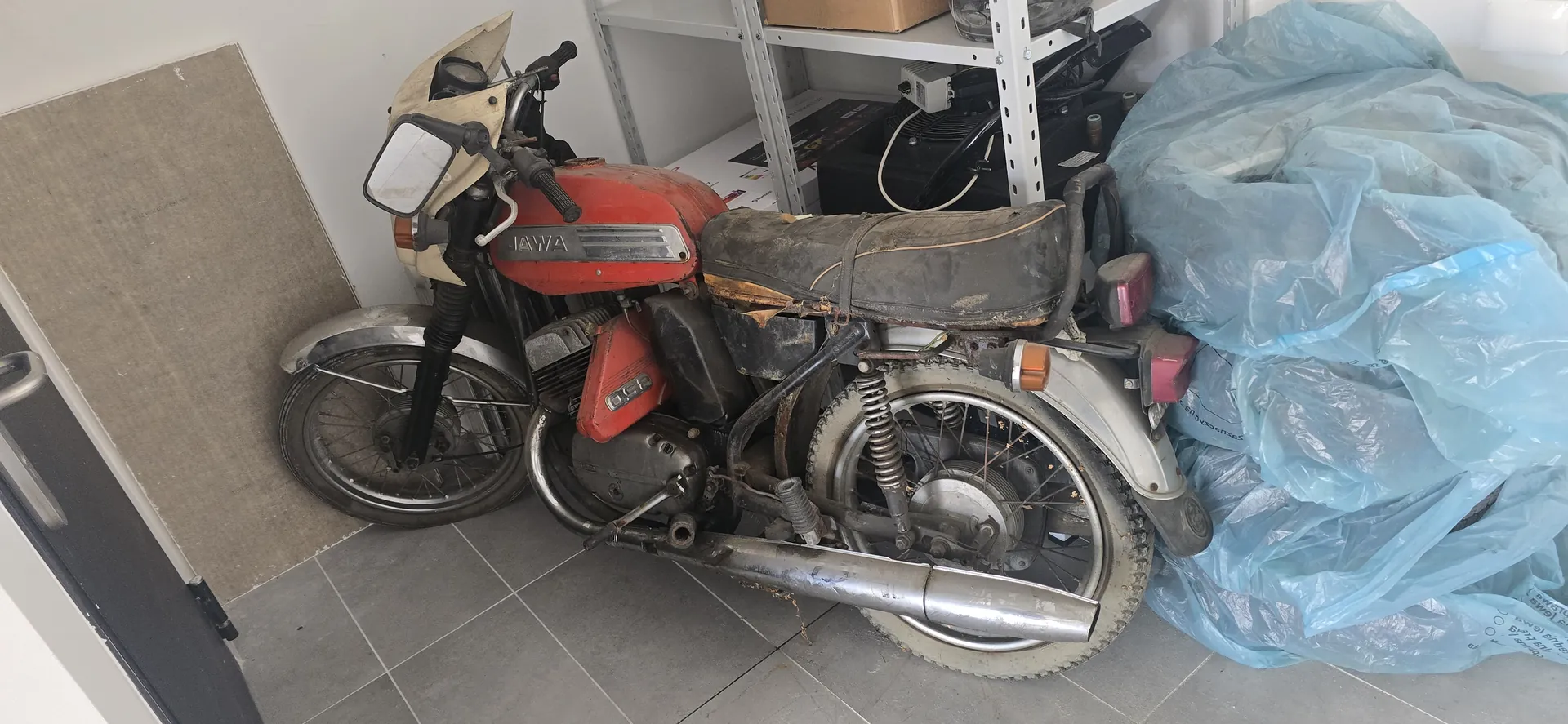 Czerwony motocykl marki JAWA z widocznym napisem "JAWA" na zbiorniku paliwa i oznaczeniem "05" na bocznej osłonie. Motocykl ma zużyte siedzenie, zarysowane elementy metalowe i opony. Znajduje się na szarych płytkach podłogowych, obok kawałka tektury opartej o białą ścianę. Po prawej stronie widoczne są przedmioty przykryte niebieską folią, a w tle metalowa półka z różnymi przedmiotami, w tym pudełkiem kartonowym i urządzeniami elektrycznymi.
