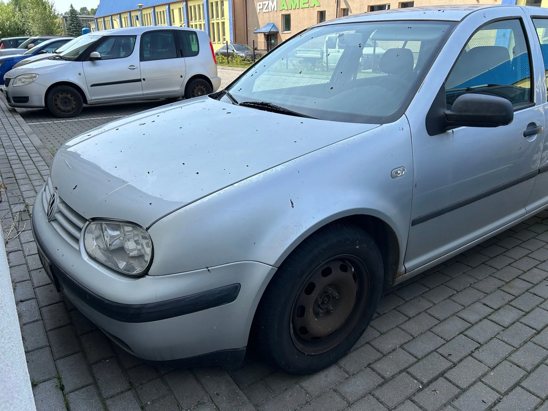Srebrny samochód osobowy marki Volkswagen Golf zaparkowany na kostce brukowej. Auto jest zakurzone i posiada liczne drobne zabrudzenia na masce. W tle widoczne są inne zaparkowane samochody oraz żółty budynek z napisem 'PZM AMEX'.