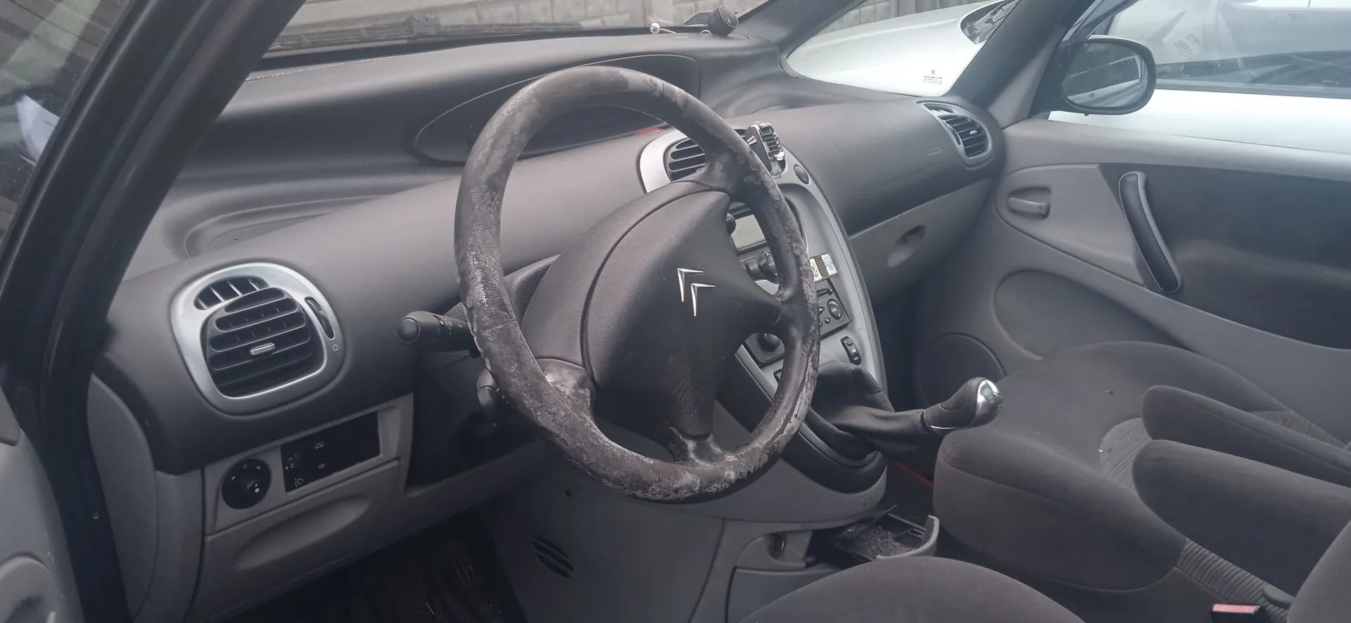 Wnętrze samochodu z perspektywy kierowcy. Widoczna jest szara deska rozdzielcza, zużyta kierownica z logo Citroën na środku, otwory nawiewów oraz ciemnoszare fotele materiałowe.