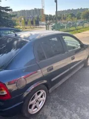 Boczny widok ciemnoniebieskiego samochodu osobowego zaparkowanego na utwardzonym placu. W tle widoczne są zielone drzewa, niskie ogrodzenie oraz wielopiętrowe budynki mieszkalne pod jasnym niebem.