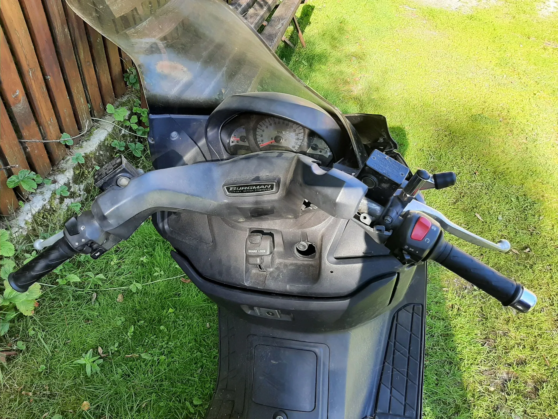 Widok z perspektywy kierowcy na kokpit czarnego skutera marki Suzuki Burgman. Widoczna jest kierownica z manetkami, klamkami hamulców, przełącznikami oraz licznik prędkości w centralnej części. Na obudowie kierownicy znajduje się napis 'BURGMAN'. Skuter stoi na trawie obok drewnianego ogrodzenia.