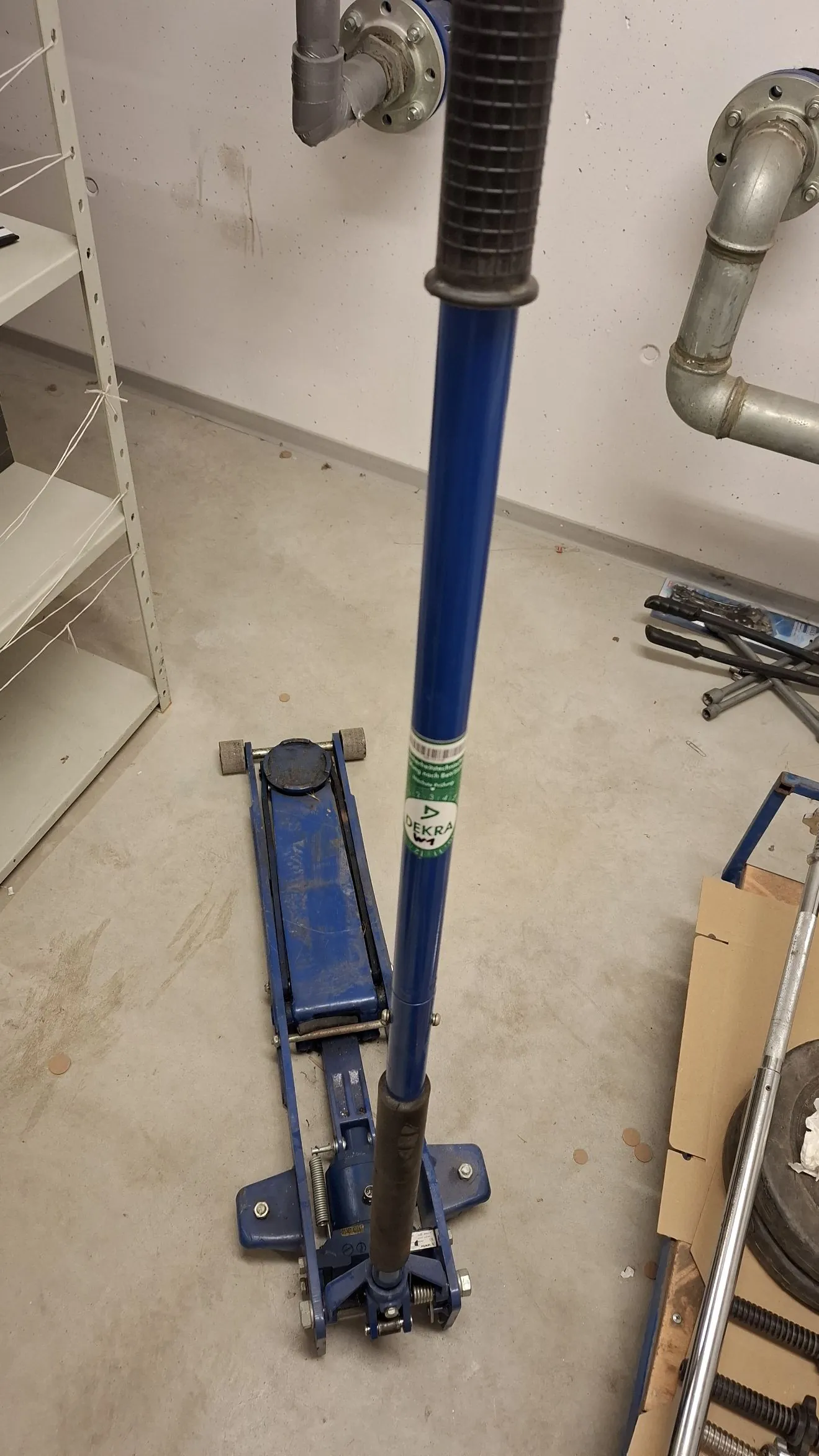 Zdjęcie przedstawiające niebieski podnośnik hydrauliczny typu żaba stojący na betonowej podłodze w pomieszczeniu gospodarczym lub warsztacie. W kadrze widoczny jest również fragment niebieskiego słupa z czarną rączką oraz rury instalacyjne w tle i biały regał po lewej stronie.