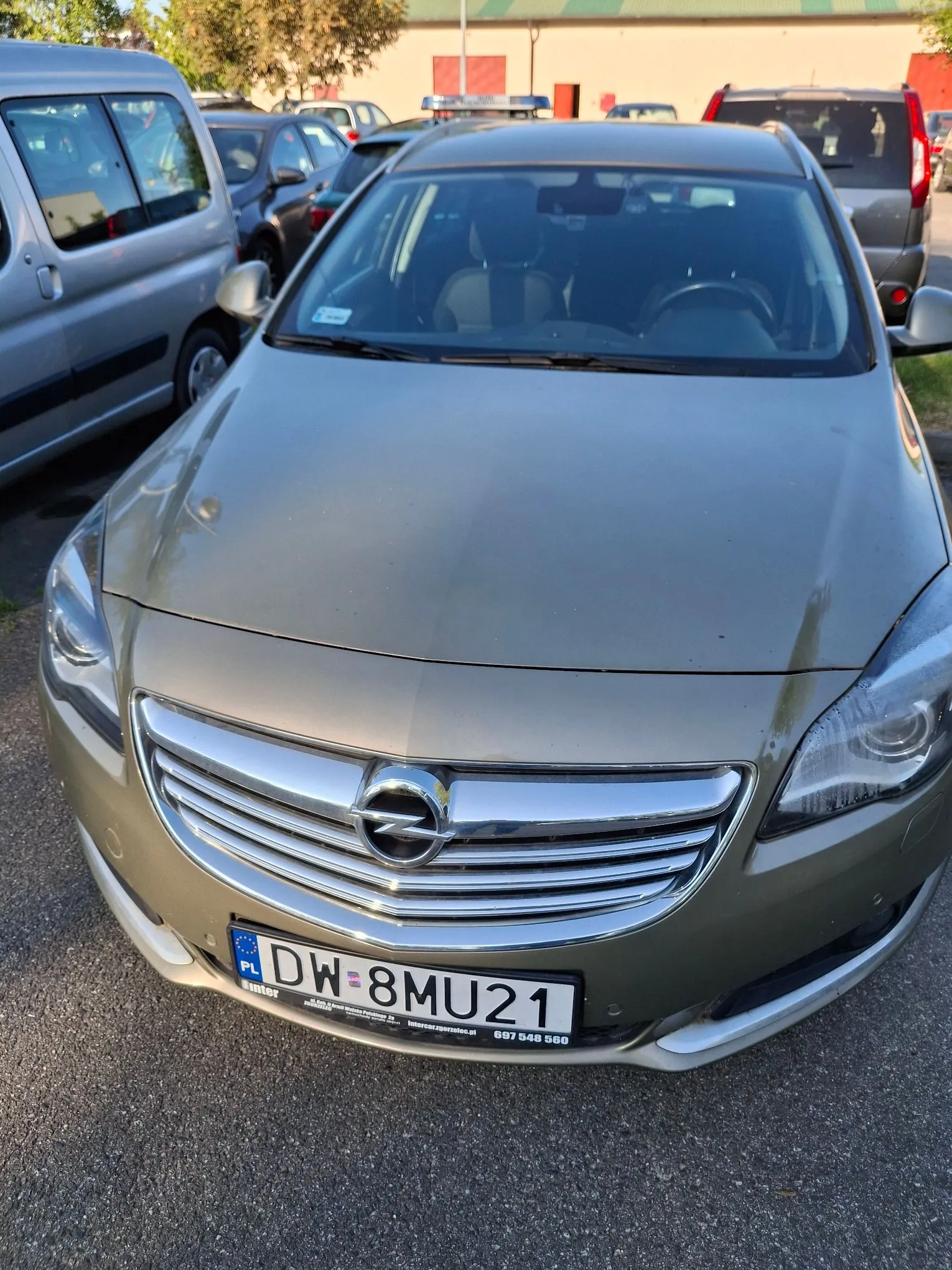 Zdjęcie przodu szarego samochodu marki Opel zaparkowanego na placu. Widoczna jest maska, grill z logo producenta oraz tablica rejestracyjna o numerze DW 8MU21. Po lewej stronie znajduje się srebrny samochód dostawczy, a w tle inne pojazdy i fragment budynku.