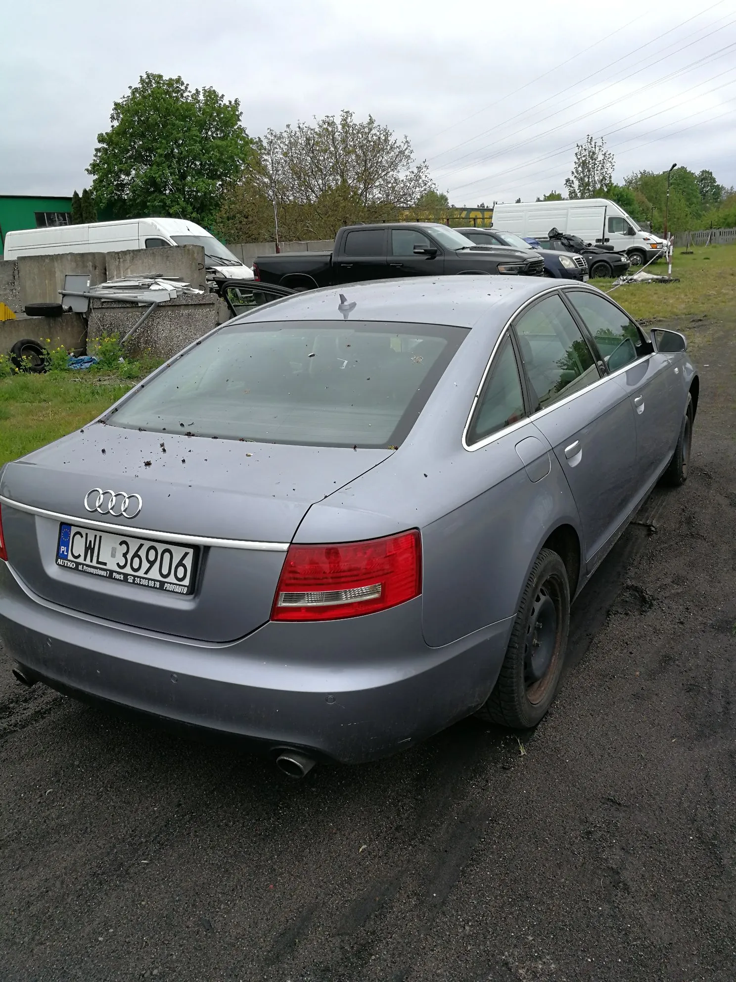 Srebrnoszary samochód marki Audi zaparkowany na piaszczystym placu. Widoczny jest tył pojazdu z polską tablicą rejestracyjną o numerze CWL 36906. W tle znajdują się inne pojazdy, w tym białe vany oraz czarny pickup, a także drzewa i betonowy mur.