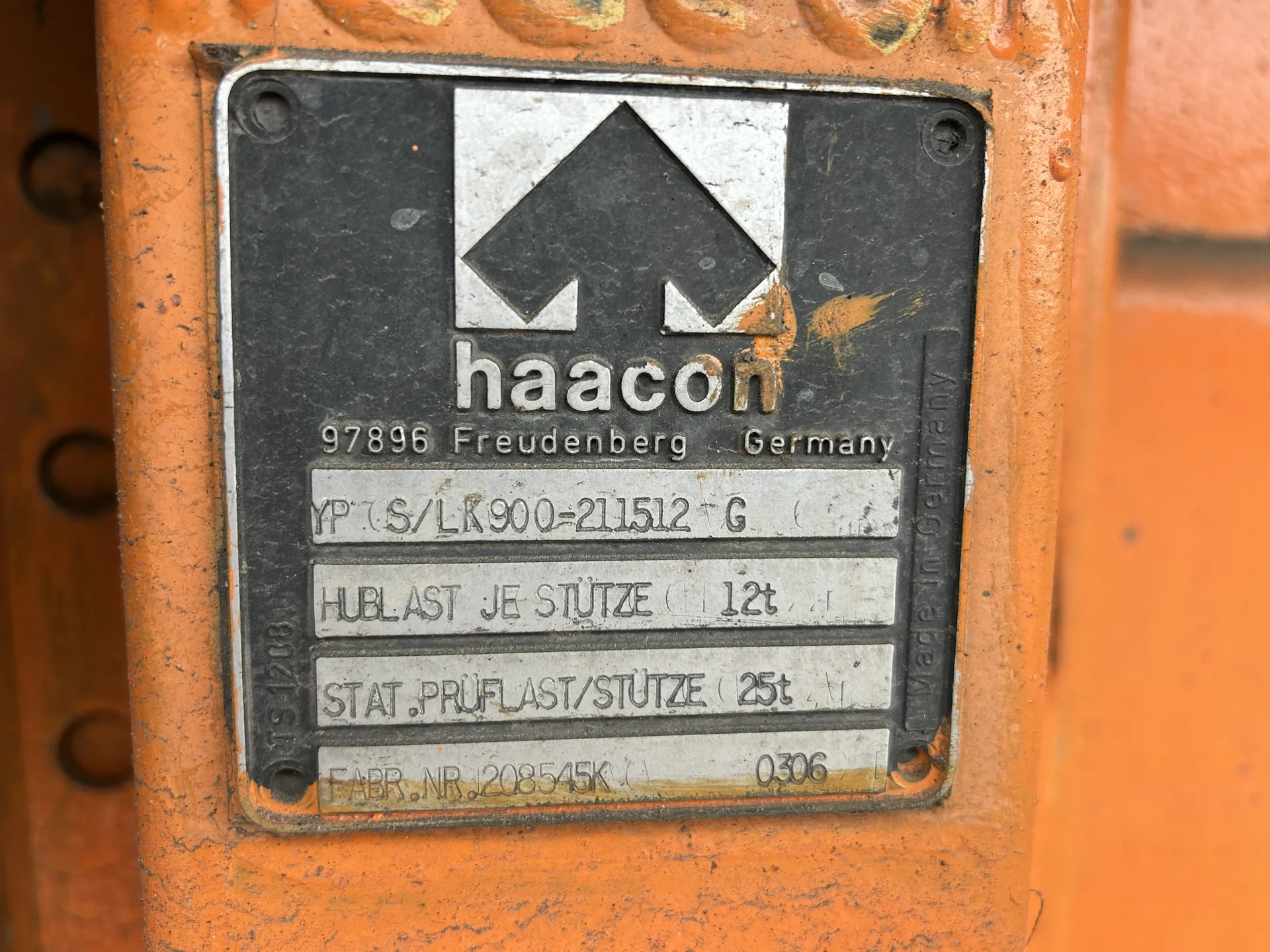Czarna metalowa tabliczka z białym logo w kształcie czarnego rombu na białym tle, poniżej napis 'haacon' w białych literach. Poniżej widoczne są napisy: '97896 Freudenberg Germany', 'YP (S/LK900-211512 G', 'HUBLAST JE STÜTZE 12t', 'STAT. PRÜFLAST/STÜTZE 25t', 'FABR. NR. 208545K 0306'. Po prawej stronie tabliczki pionowo umieszczony napis 'Made in Germany'. Tabliczka zamocowana na pomarańczowej powierzchni z widocznymi śladami zużycia i rdzy.