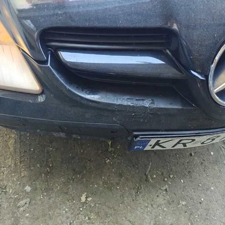 Przód ciemnego samochodu z widocznym reflektorem, czarną taśmą na zderzaku i tablicą rejestracyjną z napisem PL KR 8. Podłoga z drobnymi kamieniami i trawą.