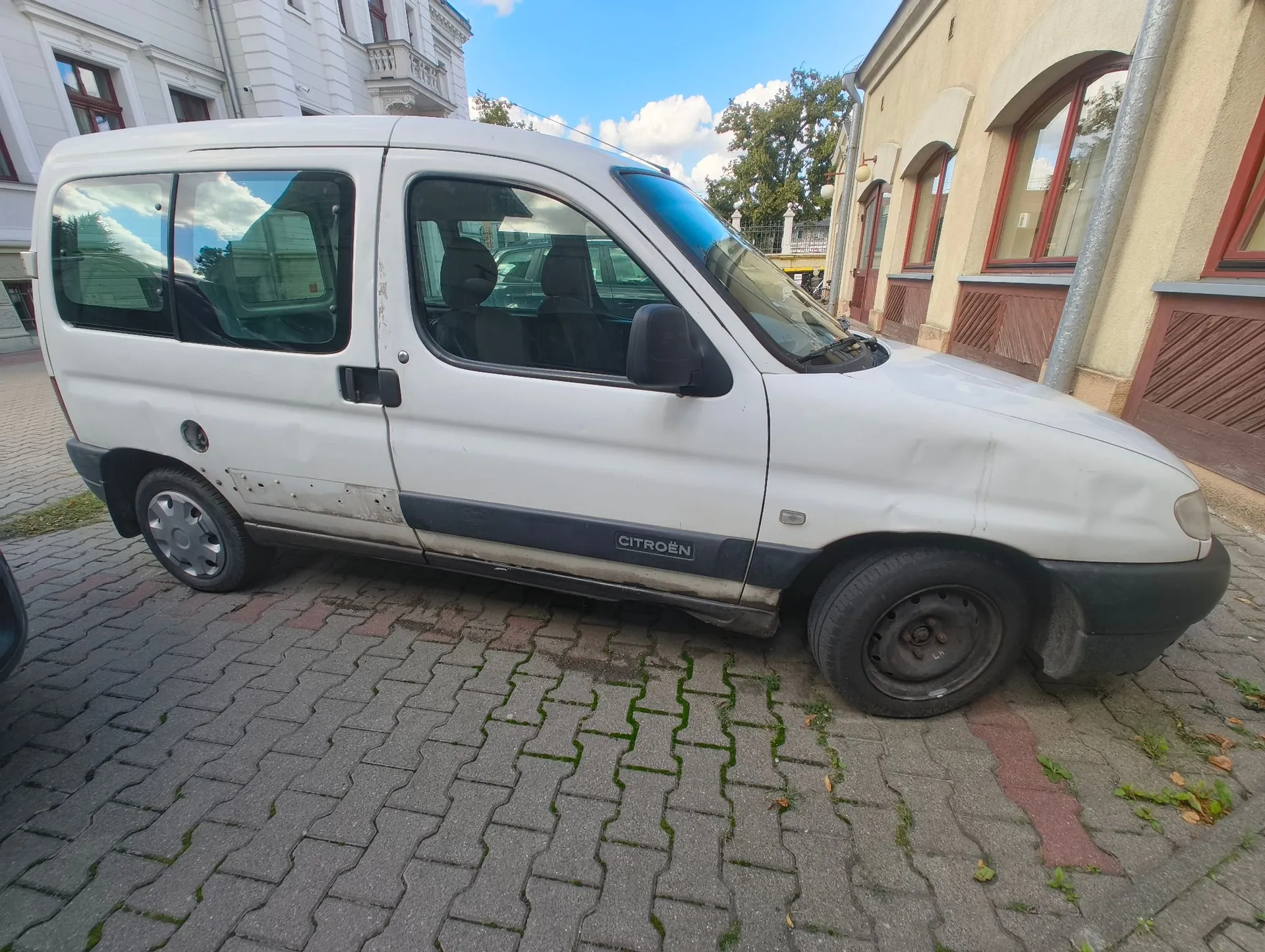 Biały samochód dostawczy marki Citroën zaparkowany na kostce brukowej przed jasnym budynkiem. Pojazd ma widoczne uszkodzenia karoserii w postaci wgnieceń na boku i z przodu. Na dolnej części drzwi widnieje napis CITROËN.