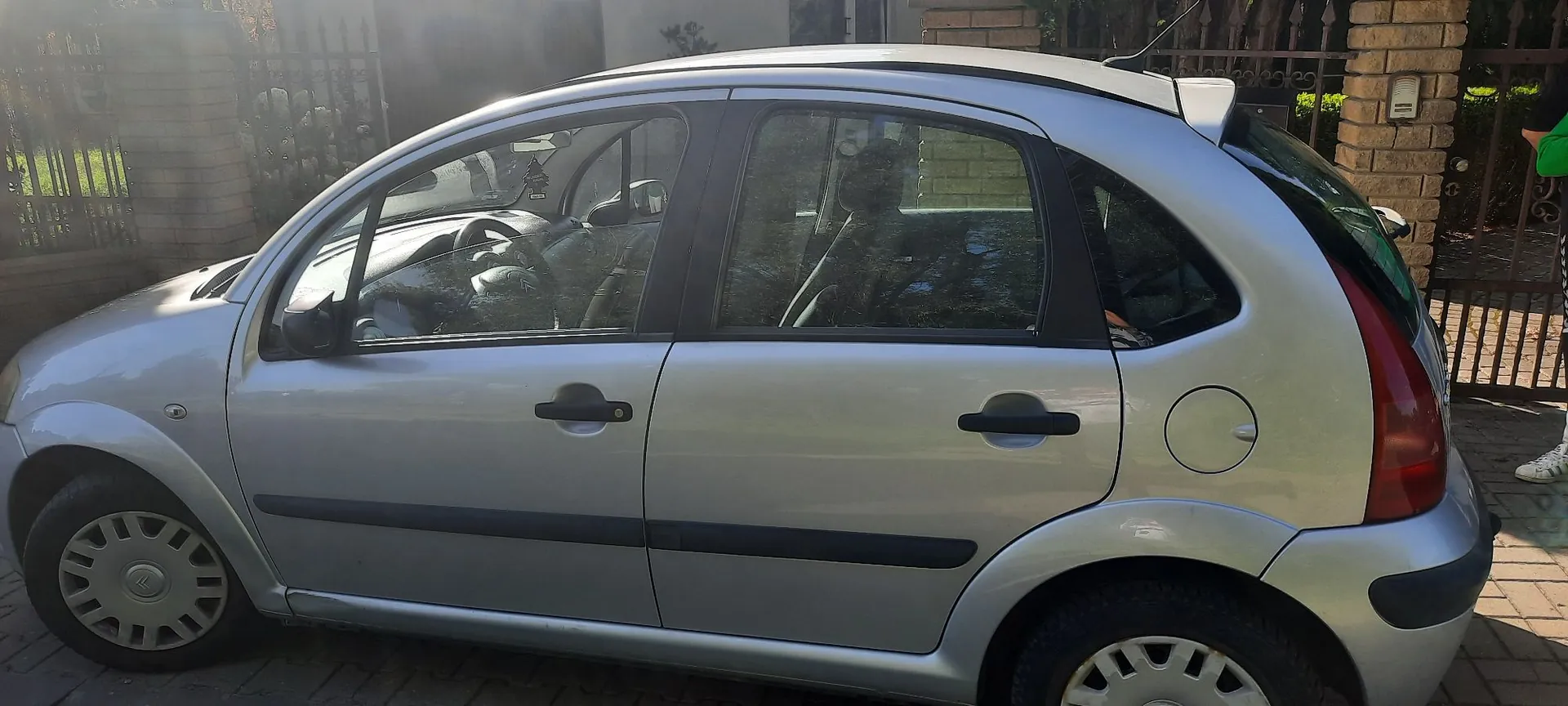 Srebrny samochód osobowy typu hatchback zaparkowany na kostce brukowej. Widok z boku, ukazujący profil pojazdu z czterema drzwiami, czarnymi listwami ochronnymi na drzwiach oraz srebrnymi felgami z kołpakami.