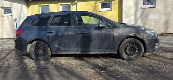 Ciemnoszary, brudny samochód typu kombi zaparkowany na podwórku z żwirową nawierzchnią. Pojazd ma zarysowane koła bez felg. W tle widoczny budynek z żółtą i beżową elewacją oraz oknami. Na ziemi i nadwoziu samochodu widoczne są cienie drzew.