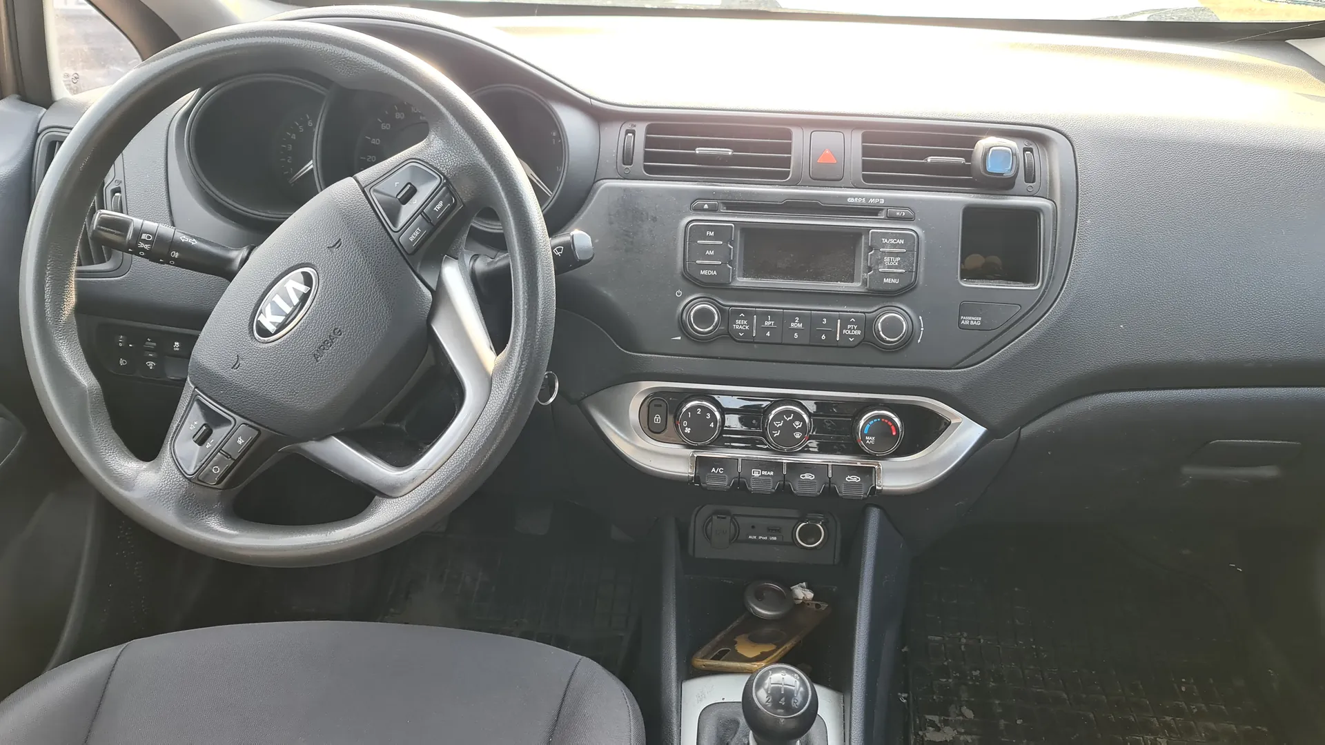 Wnętrze samochodu marki Kia z widocznym kierownicą z logo Kia, wskaźnikami prędkości i obrotomierzem, centralną konsolą z radiem oznaczonym przyciskami FM, AM, MEDIA, ekranem wyświetlacza oraz sterownikami klimatyzacji. Na środku konsoli znajduje się dźwignia zmiany biegów, a obok niej telefon w obudowie. Siedzenia są szare, a dywaniki podłogowe czarne. Wszystkie elementy są ustawione w standardowej orientacji dla wnętrza pojazdu.