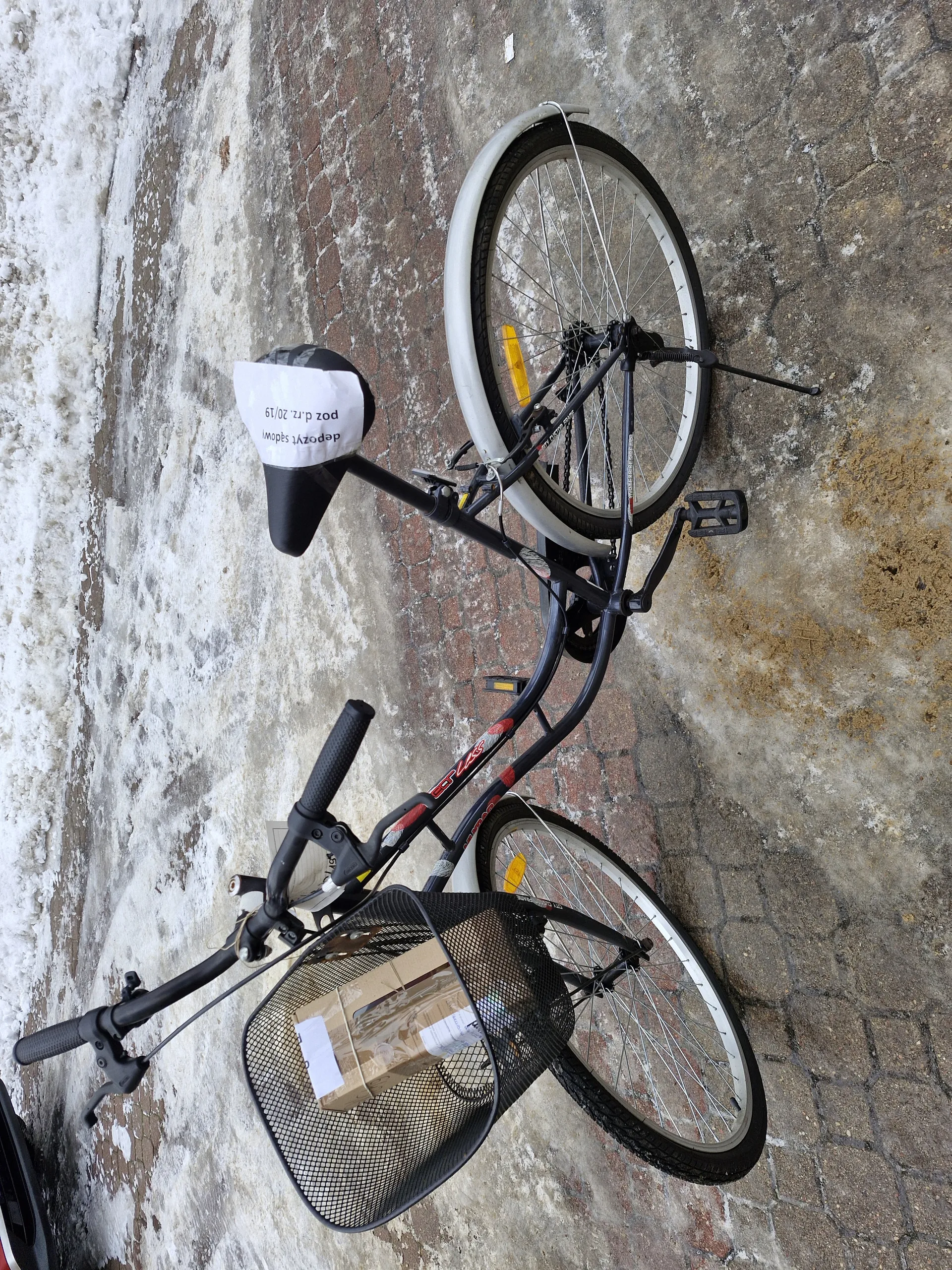 Czarny rower z przednim siatkowym koszem zawierającym kartonowe pudełko z etykietami. Rower ma białą osłonę tylnego koła, żółte reflektory oraz czarne siedzisko z białym papierem. Na papierze widoczny tekst: "depozyt 14zł" oraz "poz. d. nr 20/19". Rower zaparkowany na brukowanej powierzchni z resztkami śniegu i lodu w tle.