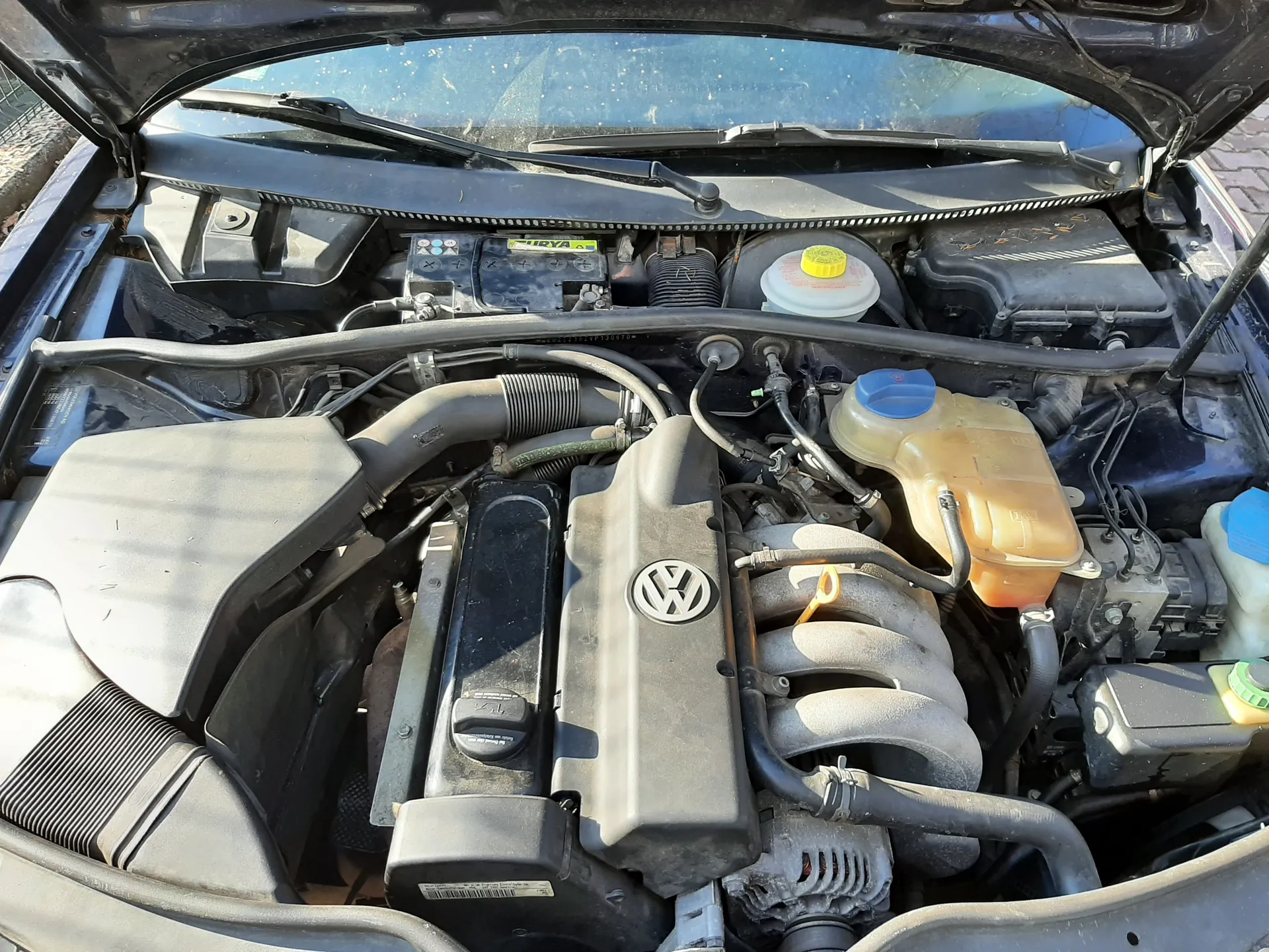 Widok na otwarty maskę samochodu z silnikiem marki Volkswagen. W centrum znajduje się pokrywa silnika z logo VW. Po lewej stronie widoczna bateria z napisem VARTA oraz czarno-białymi symbolami. Obok niej zbiornik z żółtym korkiem, prawdopodobnie na płyn chłodniczy, oraz zbiornik z niebieskim korkiem. W tle widać przednią szybę i wycieraczki. W silniku widoczne są różne przewody, rury i elementy mechaniczne.