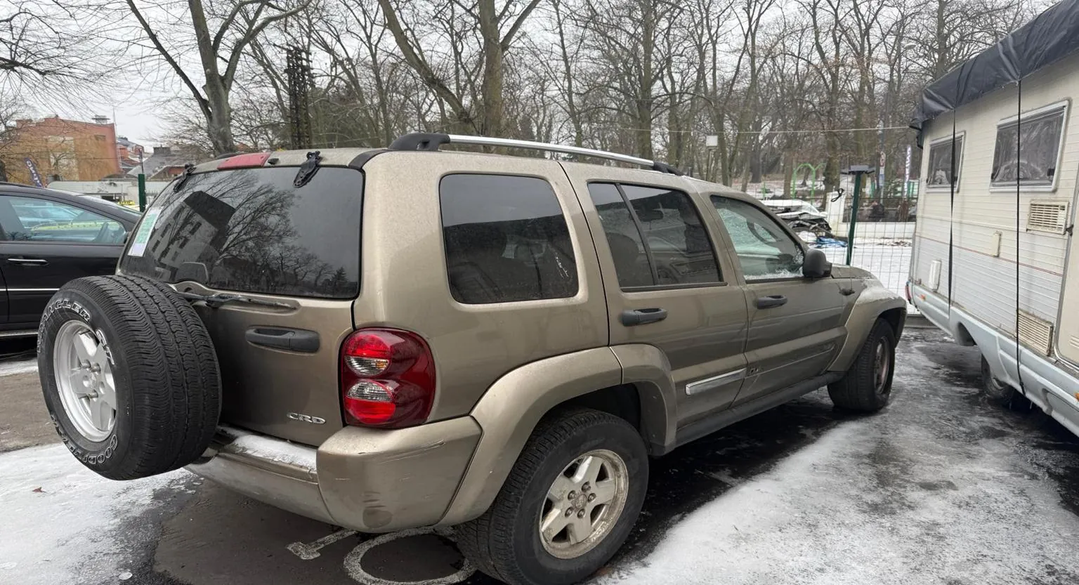 Beżowy SUV z zamontowanym na tylnej części zapasowym kołem, zaparkowany na nawierzchni częściowo pokrytej śniegiem. Po prawej stronie widoczny przyczepa kempingowa, po lewej czarny samochód. W tle drzewa bez liści oraz budynki. Na tylnej części SUV widoczny napis CRD. Opona zapasowa ma widoczny napis BFGoodrich.