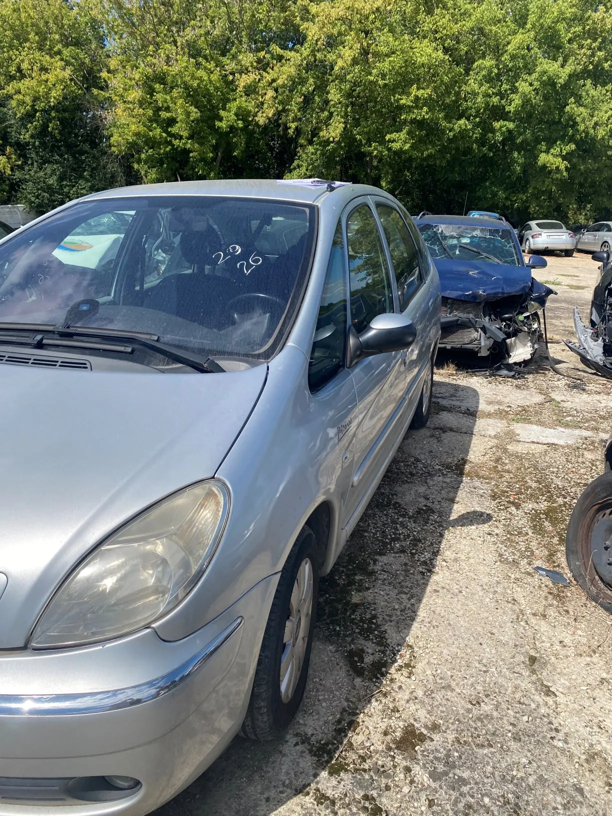 Srebrny samochód osobowy zaparkowany na nieutwardzonym placu obok innych pojazdów. Na przedniej szybie auta widnieje napis wykonany markerem: '26 L'. W tle widać zielone drzewa oraz wrak innego, ciemnego samochodu.
