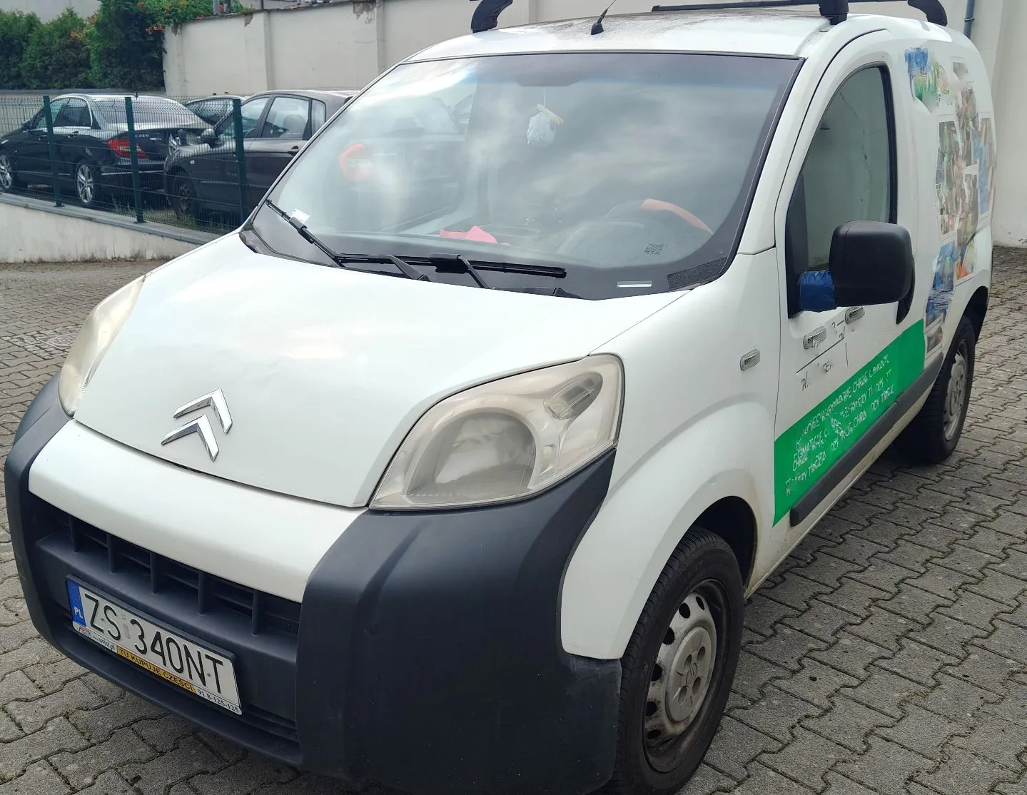 Biały samochód dostawczy marki Citroën zaparkowany na kostce brukowej. Pojazd ma czarny zderzak przedni i rejestrację ZS 340NT. Na boku auta widnieje zielony pas z tekstem oraz kolorowa grafika. W tle widoczne są inne zaparkowane samochody i biały mur.
