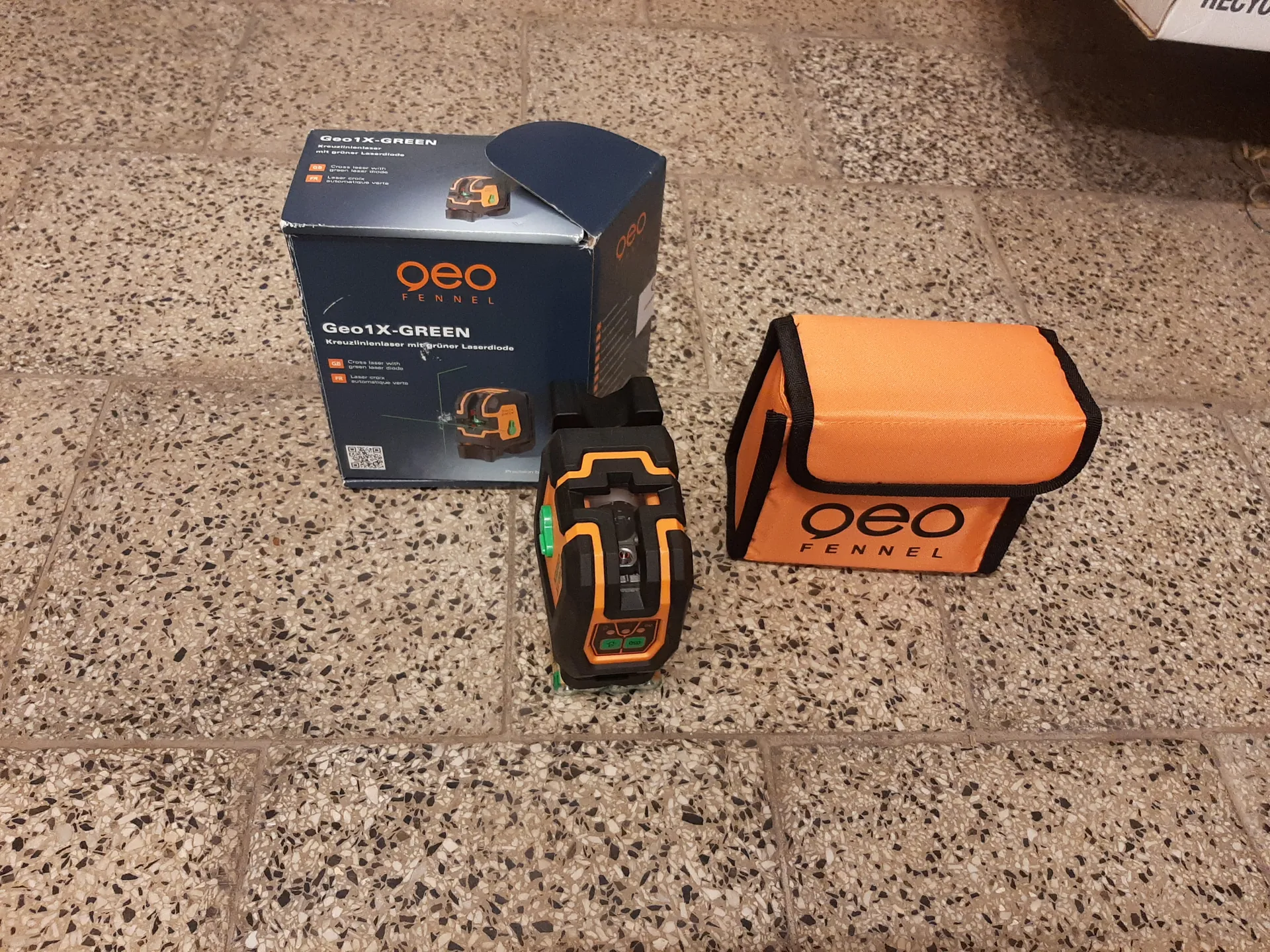 Na podłodze z kafelkami w kolorze beżowym z czarnymi i białymi kropkami znajdują się trzy przedmioty: niebieskie pudełko z napisem 'Geo1X-GREEN', 'Kreuzlinienlaser mit grüner Laserdiode' oraz logo 'geo FENNEL', czarno-pomarańczowy laser poziomnicowy z zielonymi przyciskami oraz pomarańczowa torba z czarnymi elementami i napisem 'geo FENNEL'.