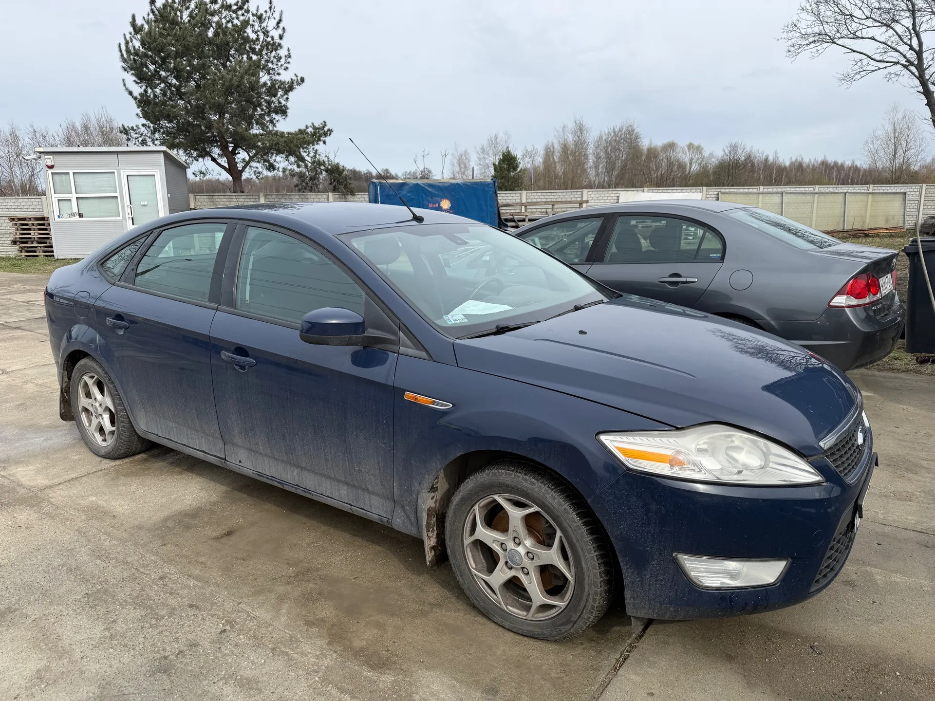 Dwa samochody osobowe zaparkowane na betonowej powierzchni. Przedni, ciemnoniebieski sedan z widocznymi śladami brudu na karoserii i felgach. Za nim szary sedan. W tle mały szary budynek z oknami i drzwiami, wysokie drzewo z zielonymi igłami, niebieska konstrukcja, betonowy płot oraz bezlistne drzewa. Niebo jest zachmurzone. Na przedniej szybie niebieskiego samochodu widoczny jest biały dokument.