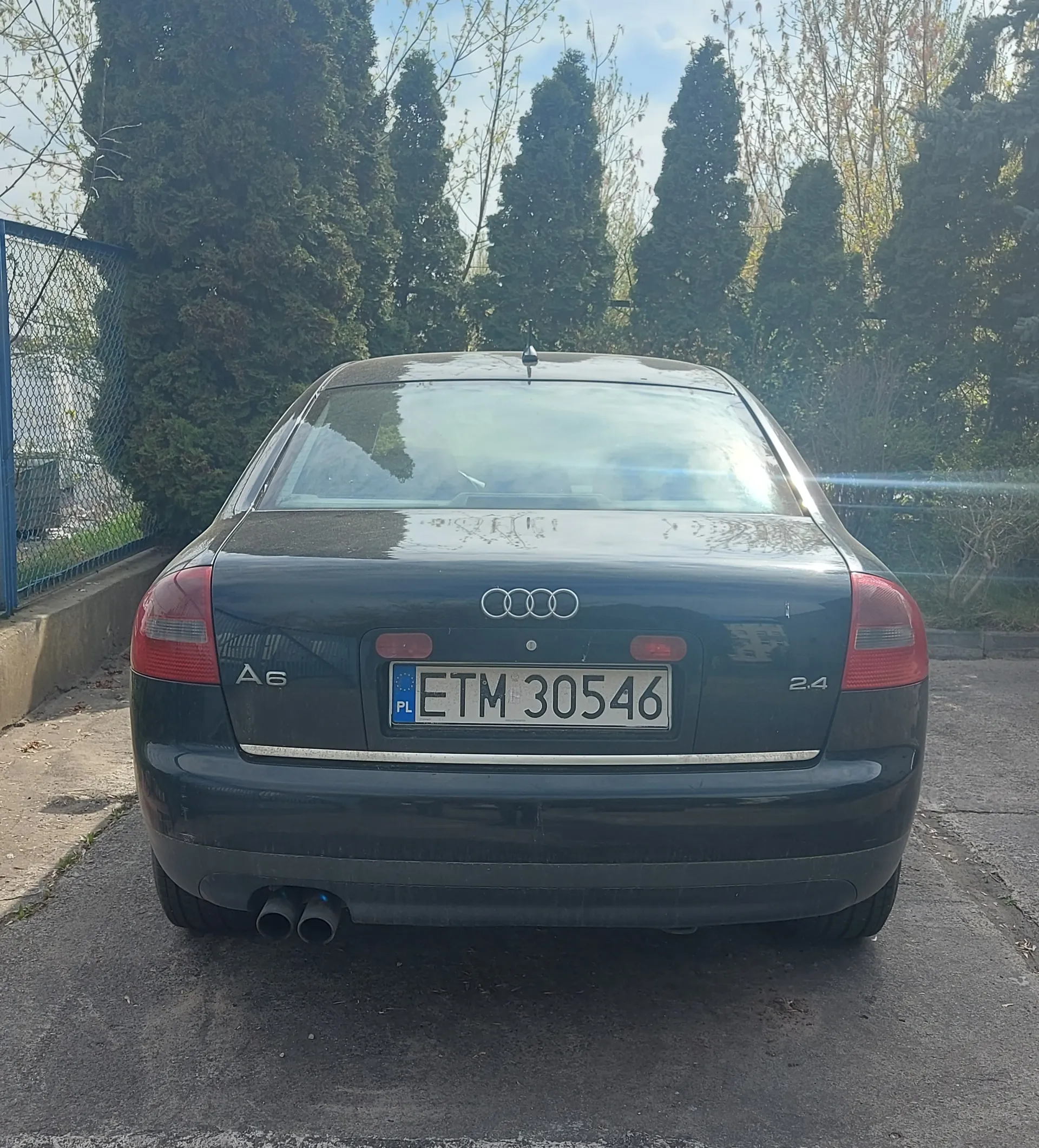 Tył ciemnozielonego samochodu Audi A6 z widocznym logo czterech pierścieni, napisem A6 po lewej stronie bagażnika i 2.4 po prawej. Tablica rejestracyjna PL ETM 30546 z niebieskim pasem po lewej stronie. Samochód stoi na asfaltowej powierzchni, w tle wysokie drzewa iglaste, niebieskie ogrodzenie siatkowe po lewej stronie oraz częściowo widoczne drzewa liściaste. Na bagażniku widoczne są czerwone światła tylnego oświetlenia i podwójne wyloty wydechowe.