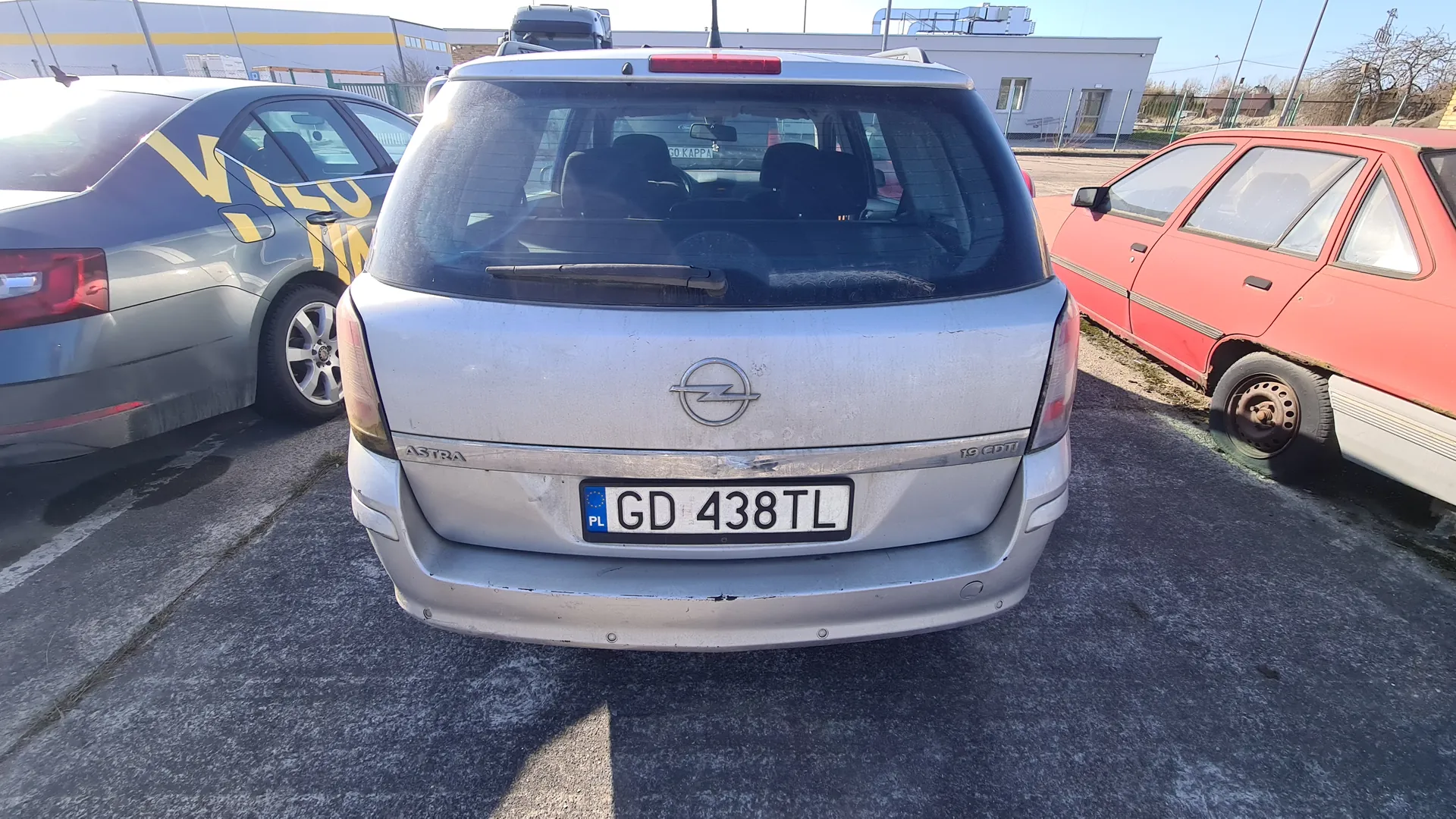 Zdjęcie przedstawia srebrny samochód Opel Astra z tyłu, z tablicą rejestracyjną GD 438TL (PL). Po lewej stronie widoczny jest szary samochód z żółtym napisem VALO, po prawej czerwony samochód z uszkodzonym kołem. Tło stanowią budynki przemysłowe i asfaltowy parking. Na tylnym szybie srebrnego Opela widoczny jest napis GO KAPPA.
