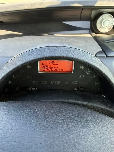 Zbliżenie na deskę rozdzielczą samochodu. W centrum znajduje się cyfrowy wyświetlacz z pomarańczowym tłem, na którym widnieją liczby: 1145.3 oraz 342052 km. Poniżej wyświetlacza znajduje się napis STOP oraz szereg kontrolek ostrzegawczych, w tym ikony oleju, akumulatora i świateł. Całość jest obudowana czarnym, plastikowym elementem kokpitu.