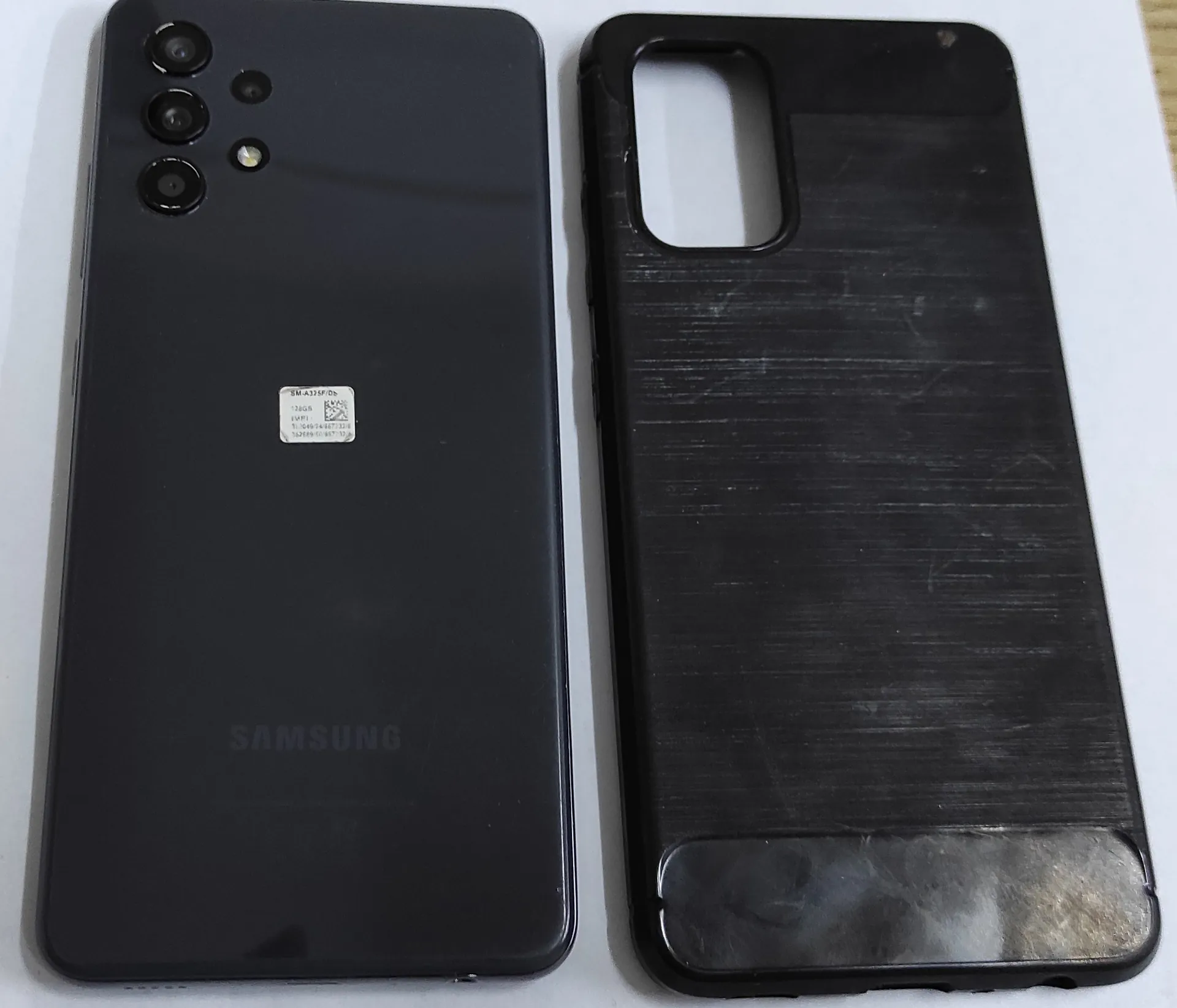 Zdjęcie przedstawiające czarny smartfon marki Samsung leżący obok czarnego, plastikowego etui ochronnego. Na tylnej obudowie telefonu widoczne są trzy obiektywy aparatu w lewym górnym rogu, biała naklejka z kodem QR i numerami seryjnymi w centralnej części oraz napis 'SAMSUNG' na dole. Etui po prawej stronie jest porysowane i zużyte.