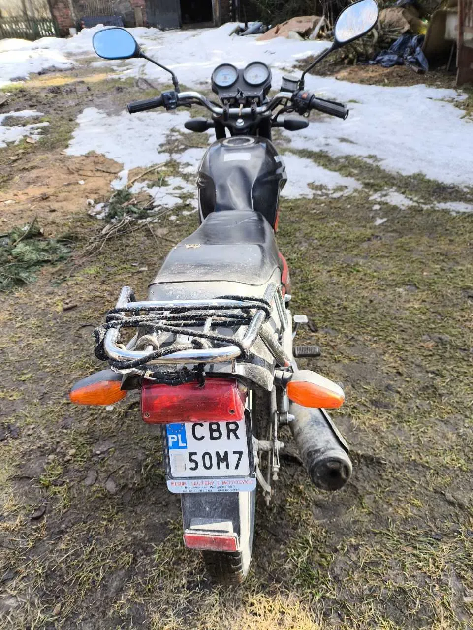 Czarny motocykl widziany od tyłu z tablicą rejestracyjną PL CBR 50M7. Motocykl ma czerwone światło tylne, pomarańczowe kierunkowskazy, dwa lusterka boczne oraz metalowy bagażnik. Tło przedstawia trawiasty teren z resztkami śniegu, budynki i różne przedmioty w oddali.