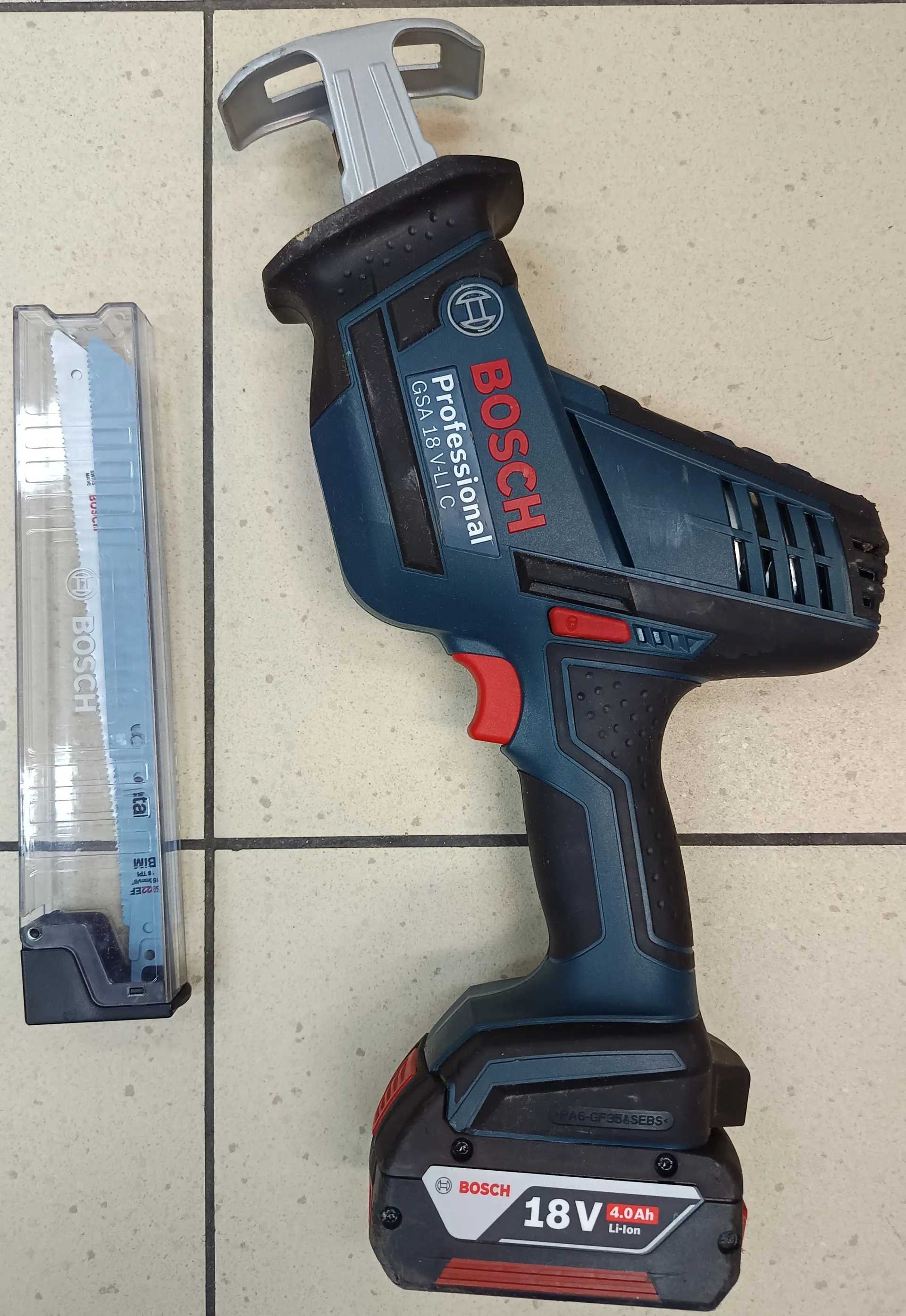 Na zdjęciu widoczna jest niebiesko-czarna wiertarka udarowa marki BOSCH Professional GSA 18V-LiC z czerwonymi akcentami. Na baterii umieszczono napisy: BOSCH, 18V, 4.0Ah, Li-Ion. Obok narzędzia leży przezroczyste etui z ostrzem BOSCH. Tło stanowi jasna płytkowa podłoga. Na narzędziu widoczne są dodatkowe oznaczenia: Professional, GSA 18V-LiC oraz logo firmy.