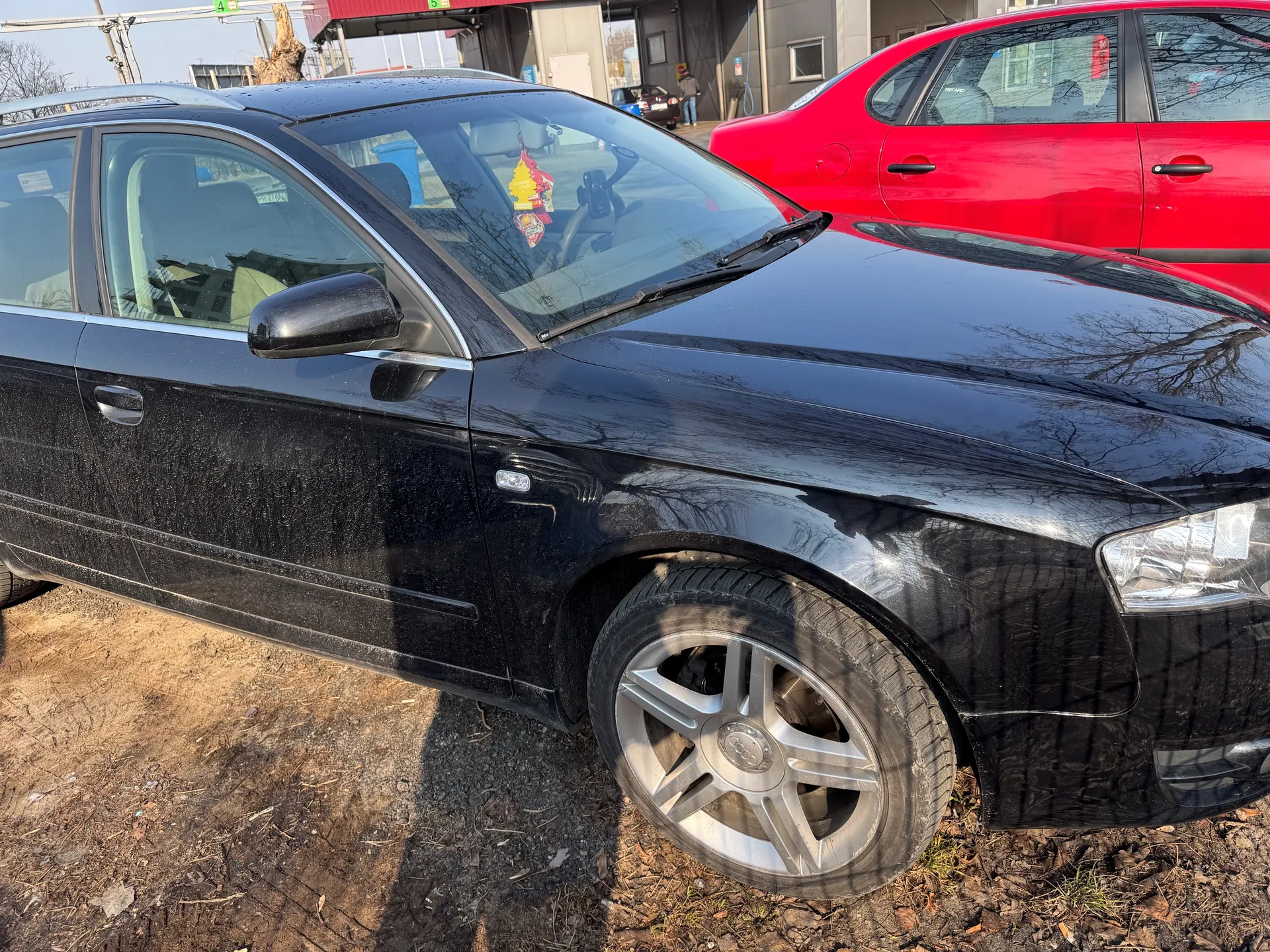 Czarny samochód Audi z przodu i boku, zaparkowany na podłożu z ziemią i żwirem. W tle czerwony samochód osobowy oraz budynek z czerwonym daszkiem. Na przedniej szybie czarnego samochodu widoczny żółty zapachowy dekorator. Koło przednie z srebrną felgą z logo Audi. Tło zawiera suche drzewa i jasne niebo.