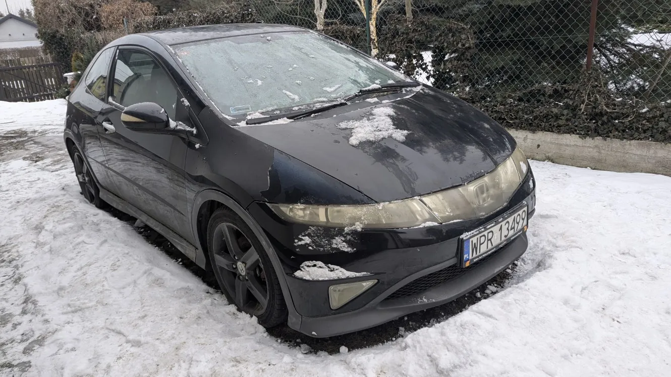 Czarny samochód marki Honda zaparkowany na śniegu. Na masce i przedniej szybie widoczne są resztki śniegu. Tablica rejestracyjna z napisem PL WPR 13499. W tle siatka ogrodzeniowa, krzewy i dom. Podłoga pokryta śniegiem.
